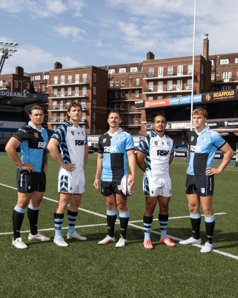 A group of five rugby players standing on a field, wearing different team jerseys. The players are posing together with a rugby ball, showcasing their team colors and styles.