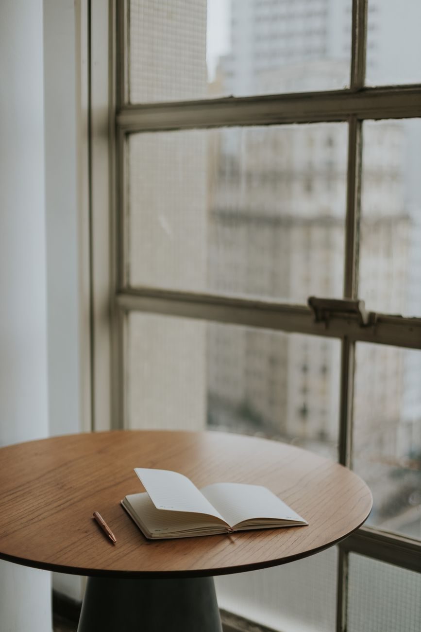 An open notebook and pencil on a wooden table beside a window