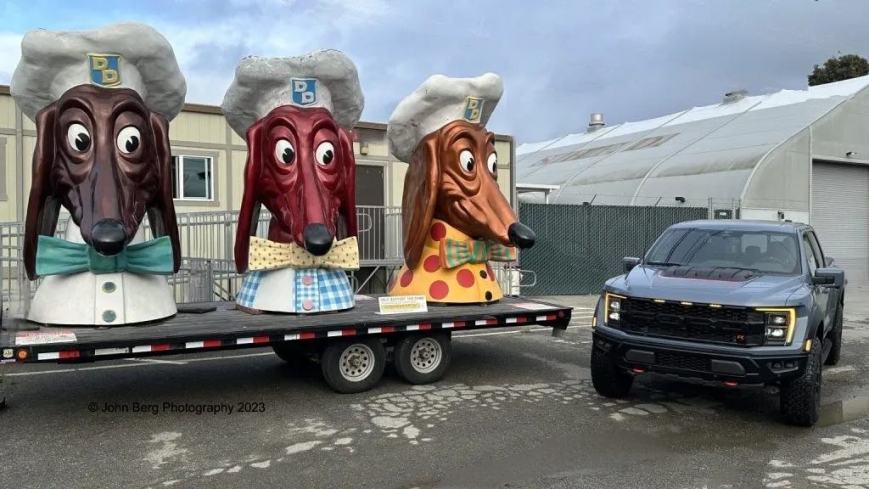 Three large dog-shaped statues in chef hats are displayed on a trailer ( mascot of the hot dog chain Doggie Diner ) alongside a gray F-150 Raptor R pickup truck. The statues have distinct color patterns, including a brown and white base. The background features a building and a greenhouse.
