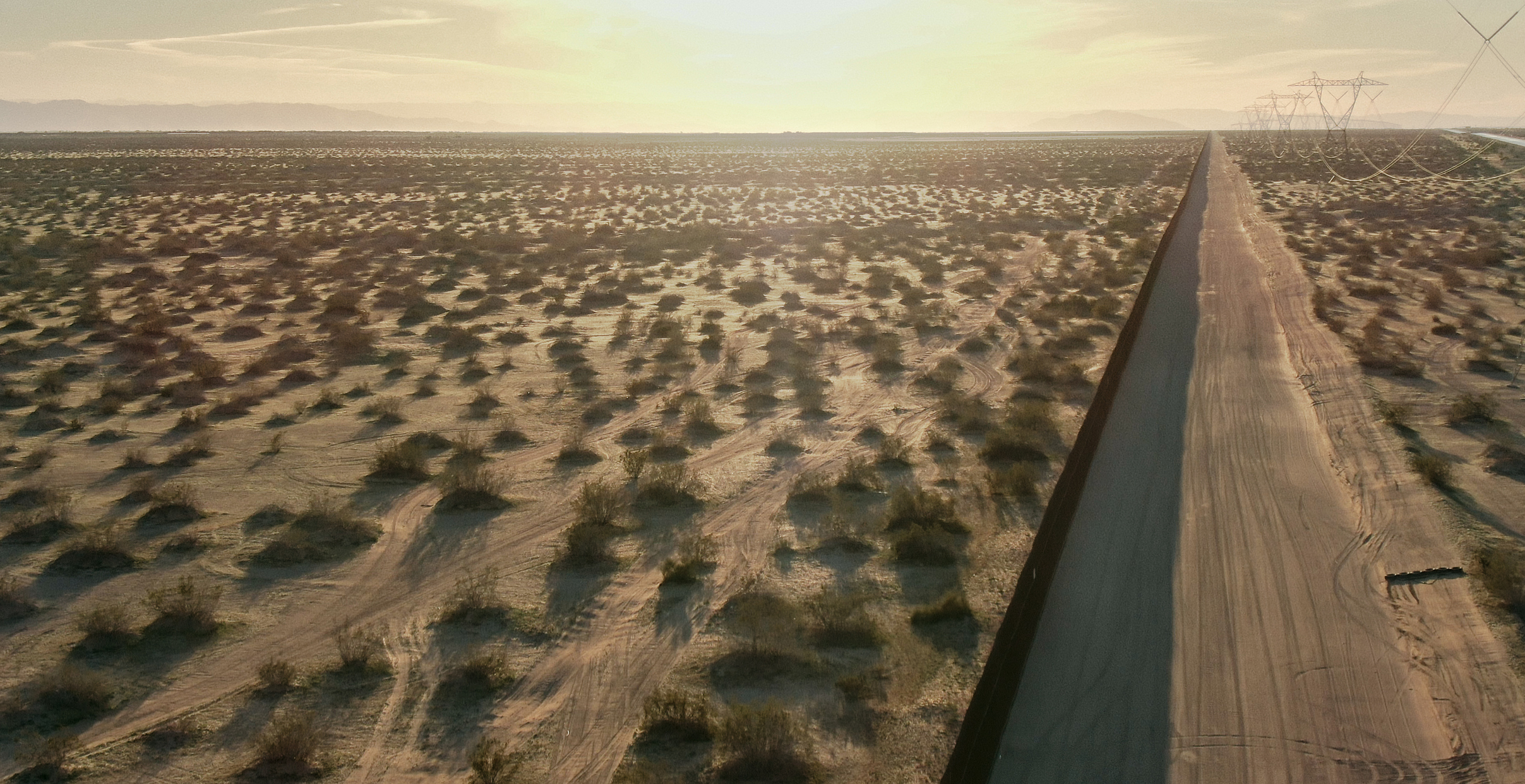 Aerial Drone Shot of a Dirt Road Running Parallel to the Steel-Slat Border Wall between Mexico and the United States on a Sunny Afternoon in the California/Mexican Desert with Distant Mountains in the Background