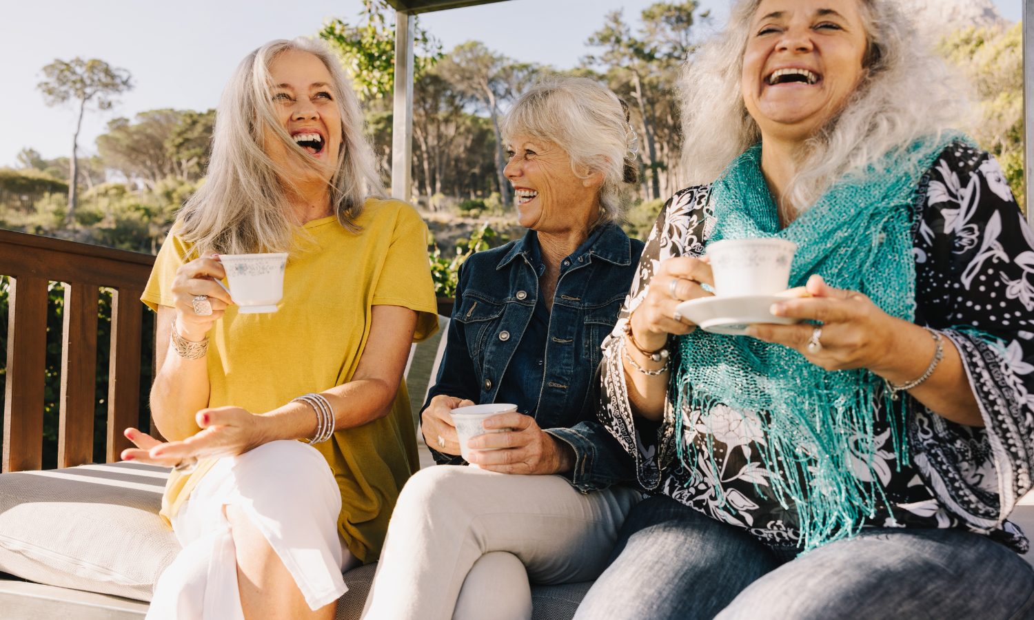 Three older women sitting together outdoors, laughing and enjoying tea from floral teacups on a sunny patio.