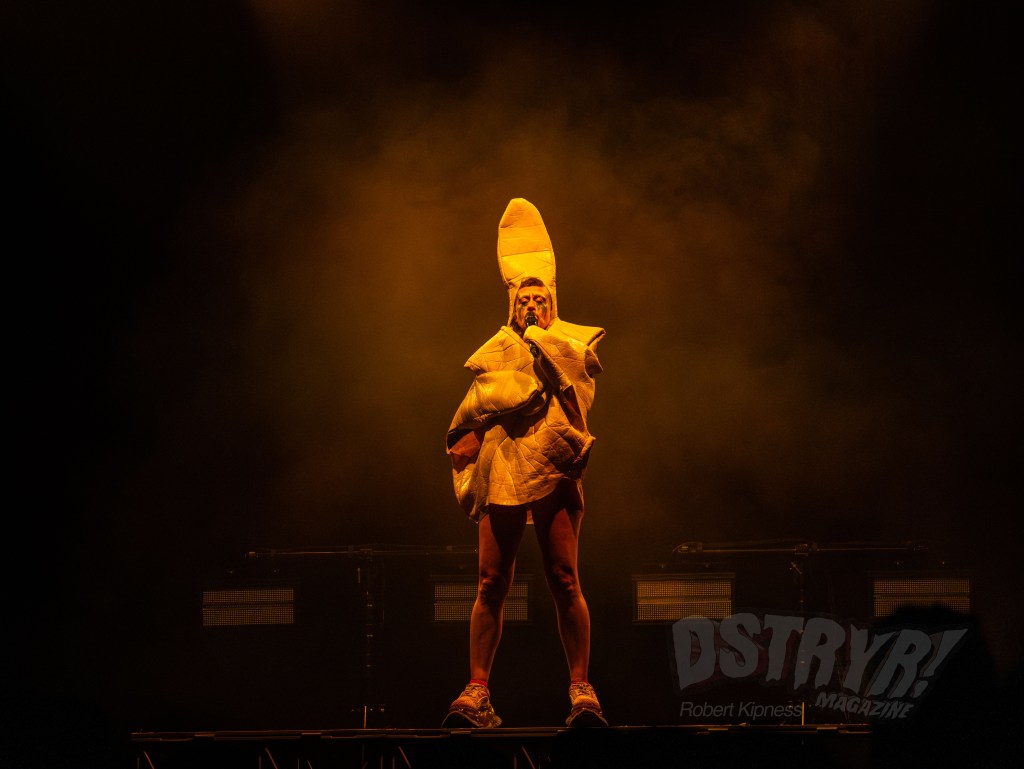 A performer dressed in a unique costume stands on stage, illuminated by dramatic lighting in a dark setting.