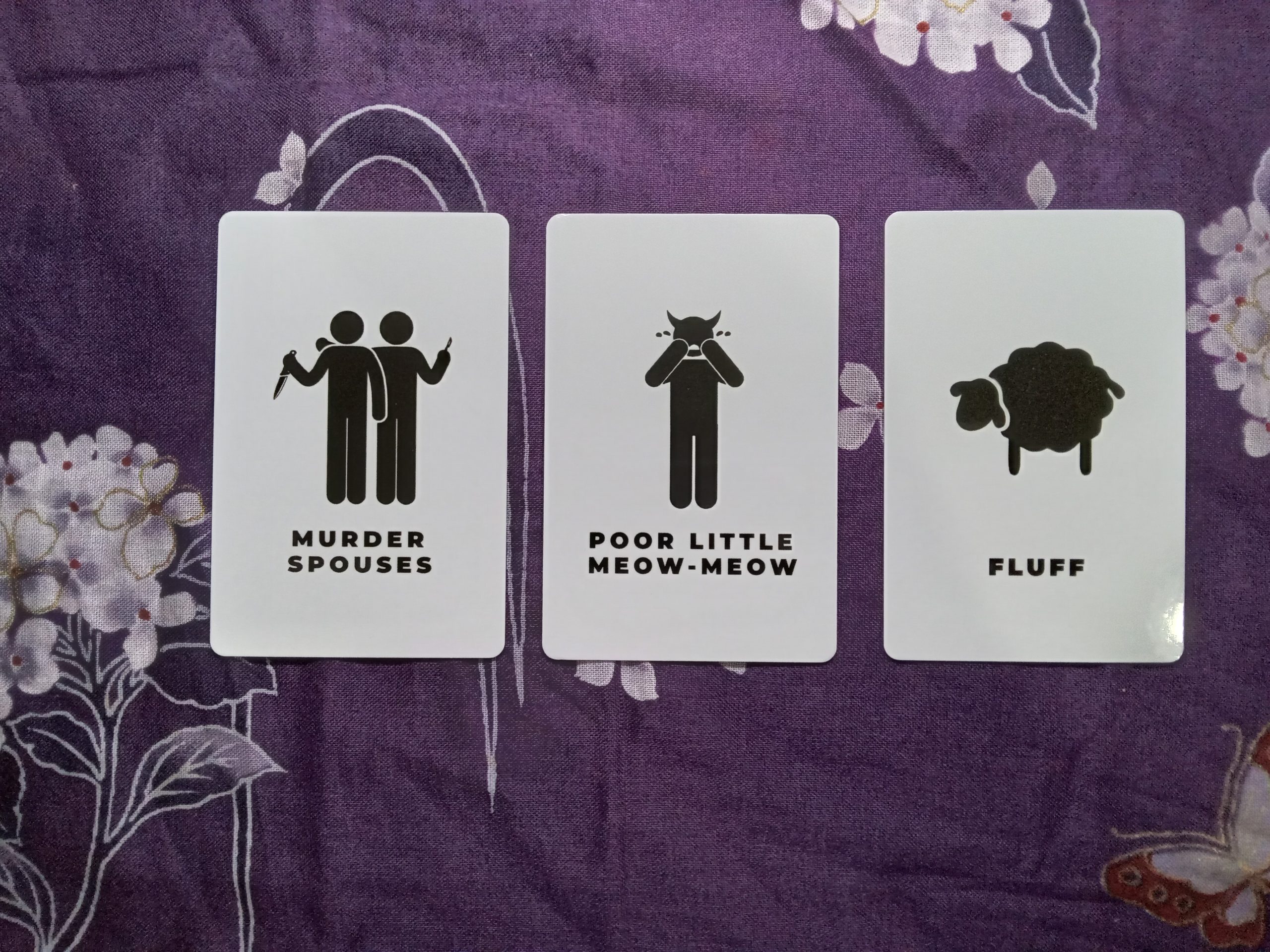 Three cards are laid on a purple fabric with white floral motif. All the cards are right-side-up. The leftmost card reads “Murder Spouses” with an image of two stick figures; one is stood behind the other holding a scalpel while the one in front has their left forearm half-raised with a knife in their hand. The middle card reads “Poor Little Meow-Meow” with an image of a standing figure with their hands raised to their face, tears coming from their eyes; they have devil horns on their head. The rightmost card reads “Fluff” with an image of a sheep.