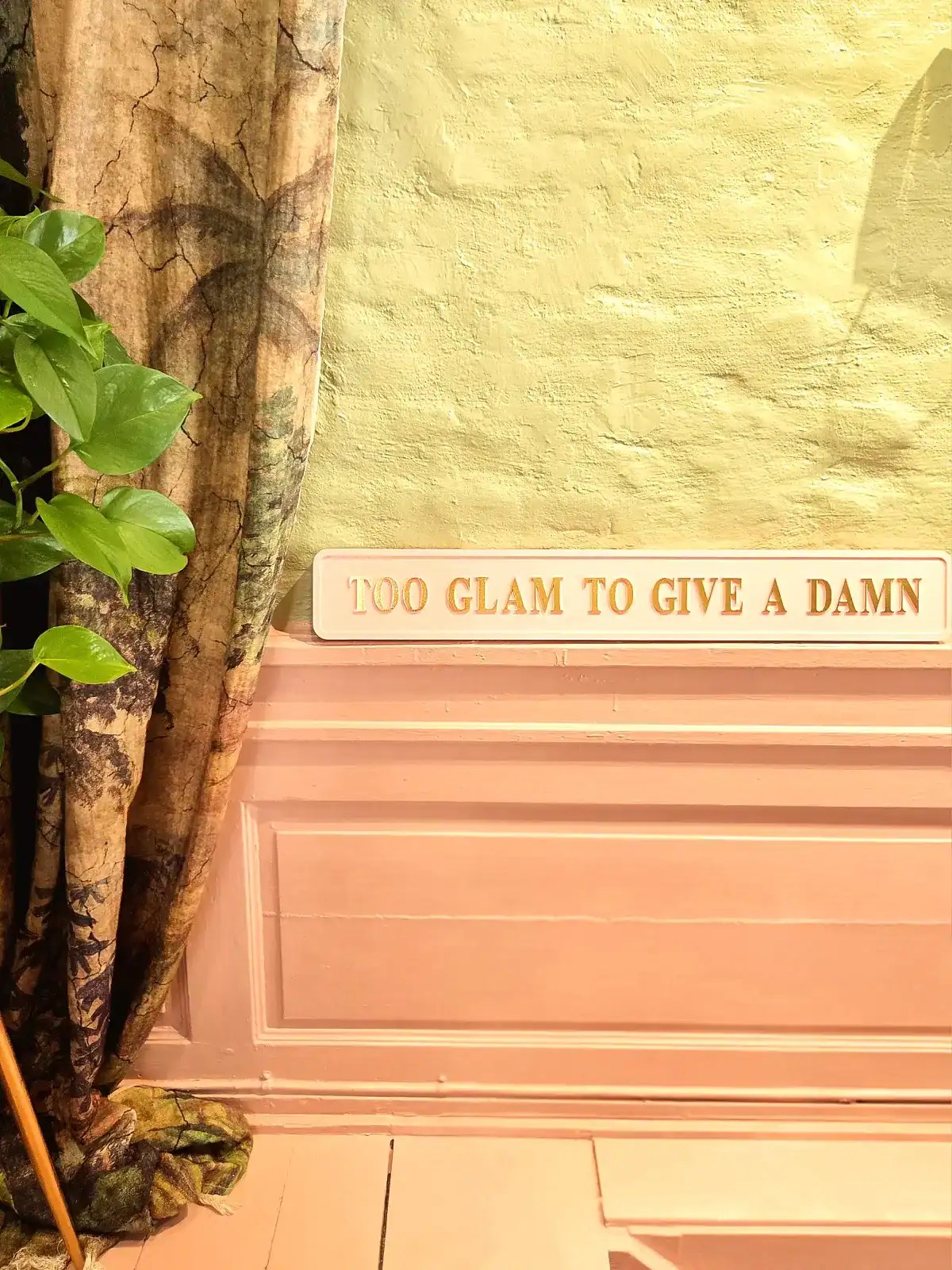 Dekorativ bogstøtte med teksten 'Glam to give a damn' i guld på lyserødt baggrund.
