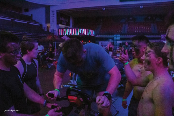 A group of individuals participating in a fitness event, focused on a person using an exercise machine. Some attendees are offering encouragement and support, with visible expressions of concentration and excitement in a dimly lit arena.
