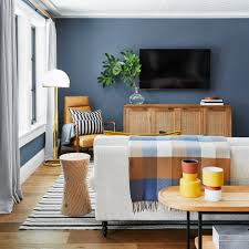 Blue paint colors for living room: Contemporary living space with medium blue walls, wooden media console, and modern decor featuring yellow accents and striped textiles for a fresh, casual look.