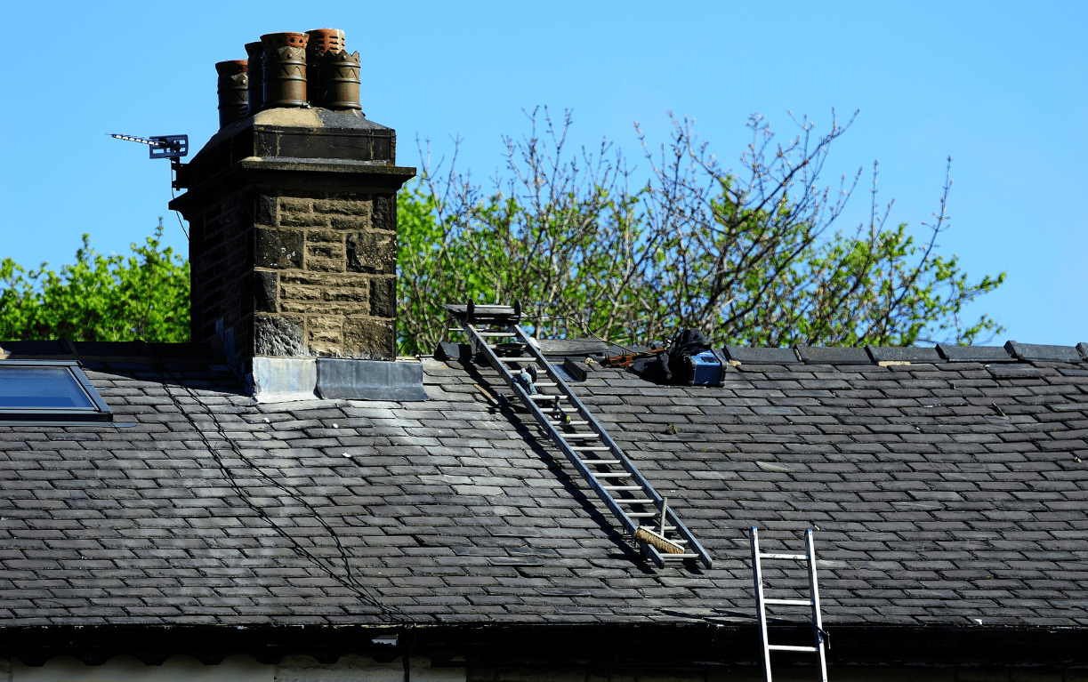 A technician using ladders and tools to inspect and repair a stone chimney on a shingled roof under clear blue skies.