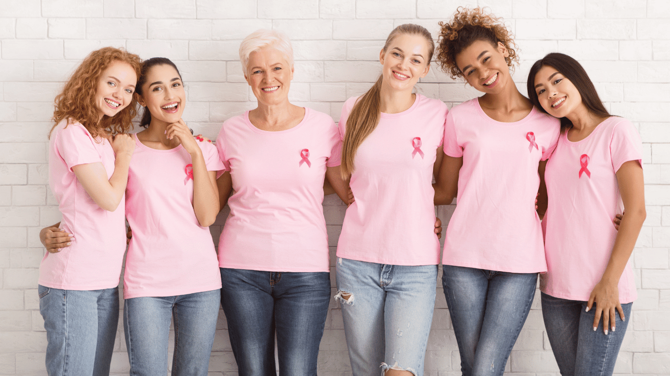 Six women standing together in pink shirts with ribbons supporting breast cancer awareness.