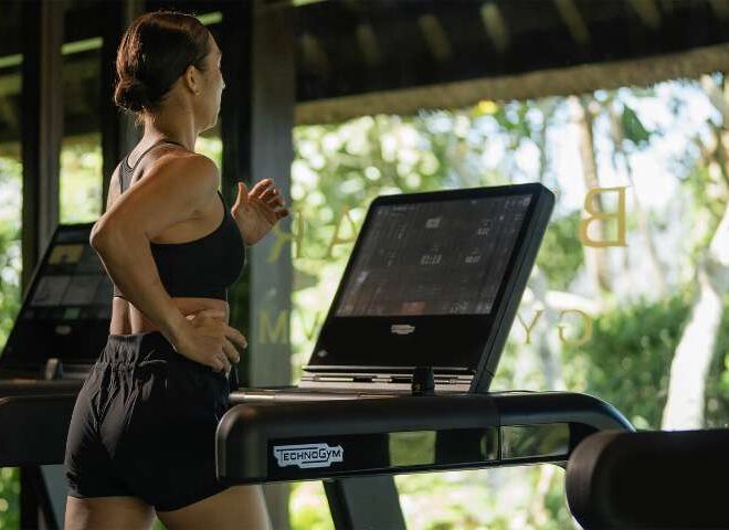 Woman running on a Technogym treadmill in the open-air gym, greenery visible through the windows