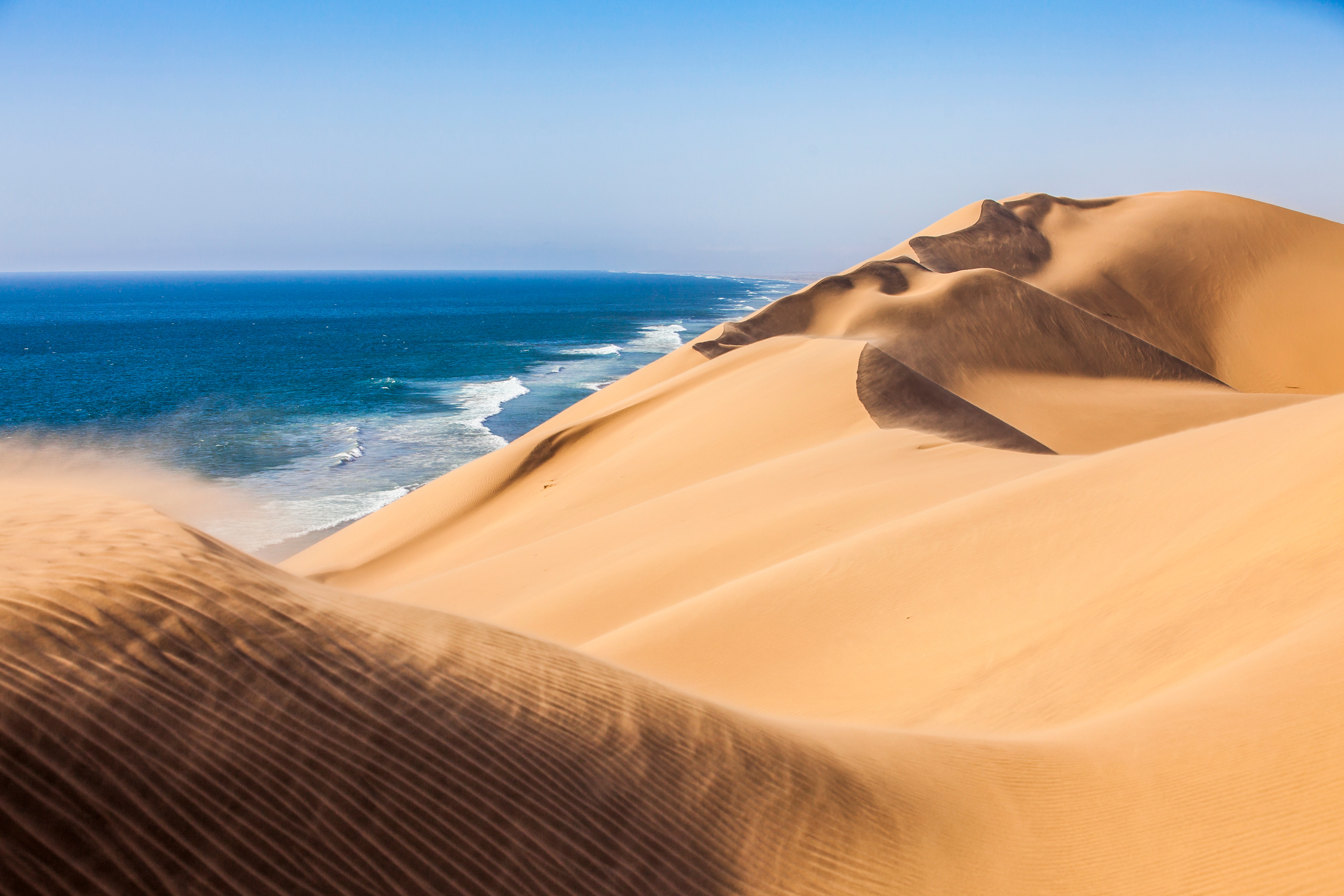 Towering sand dunes of the Namib Desert meet the Atlantic Ocean coastline, with wind-blown sand streaming off the ridges