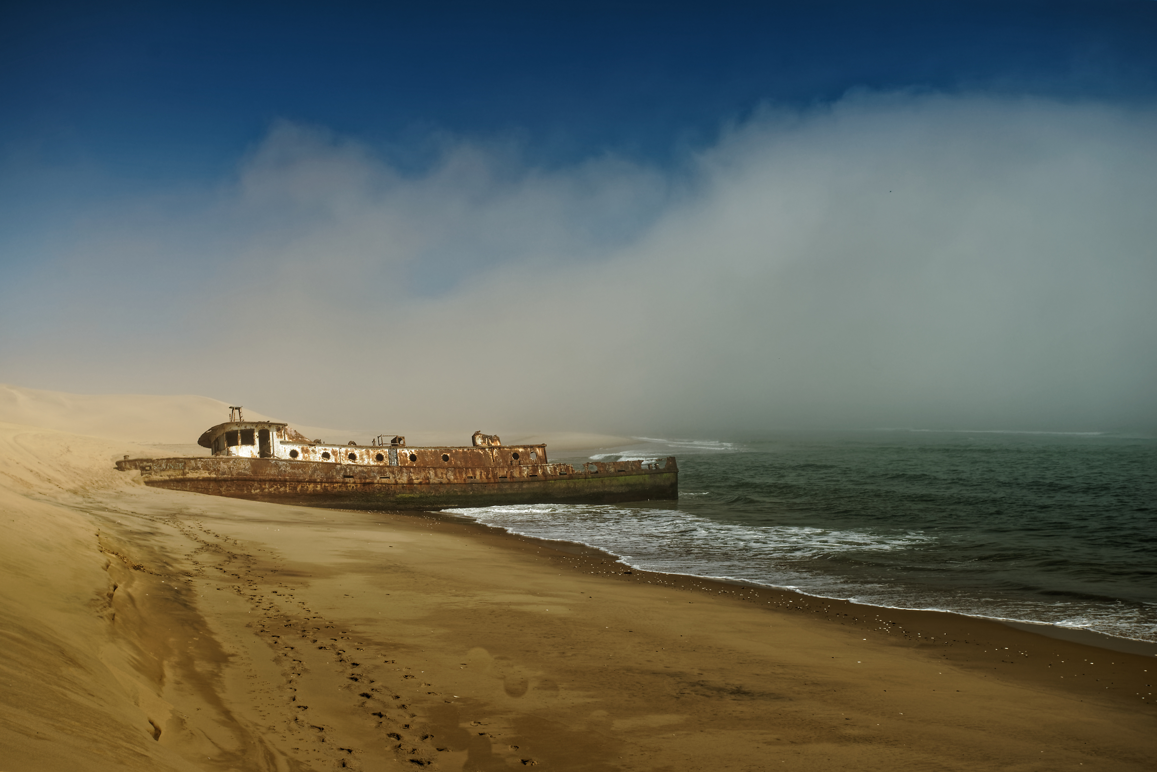 Rusted shipwreck on the Skeleton Coast of Namibia, half buried in sand with sea fog rolling in over the ocean behind it