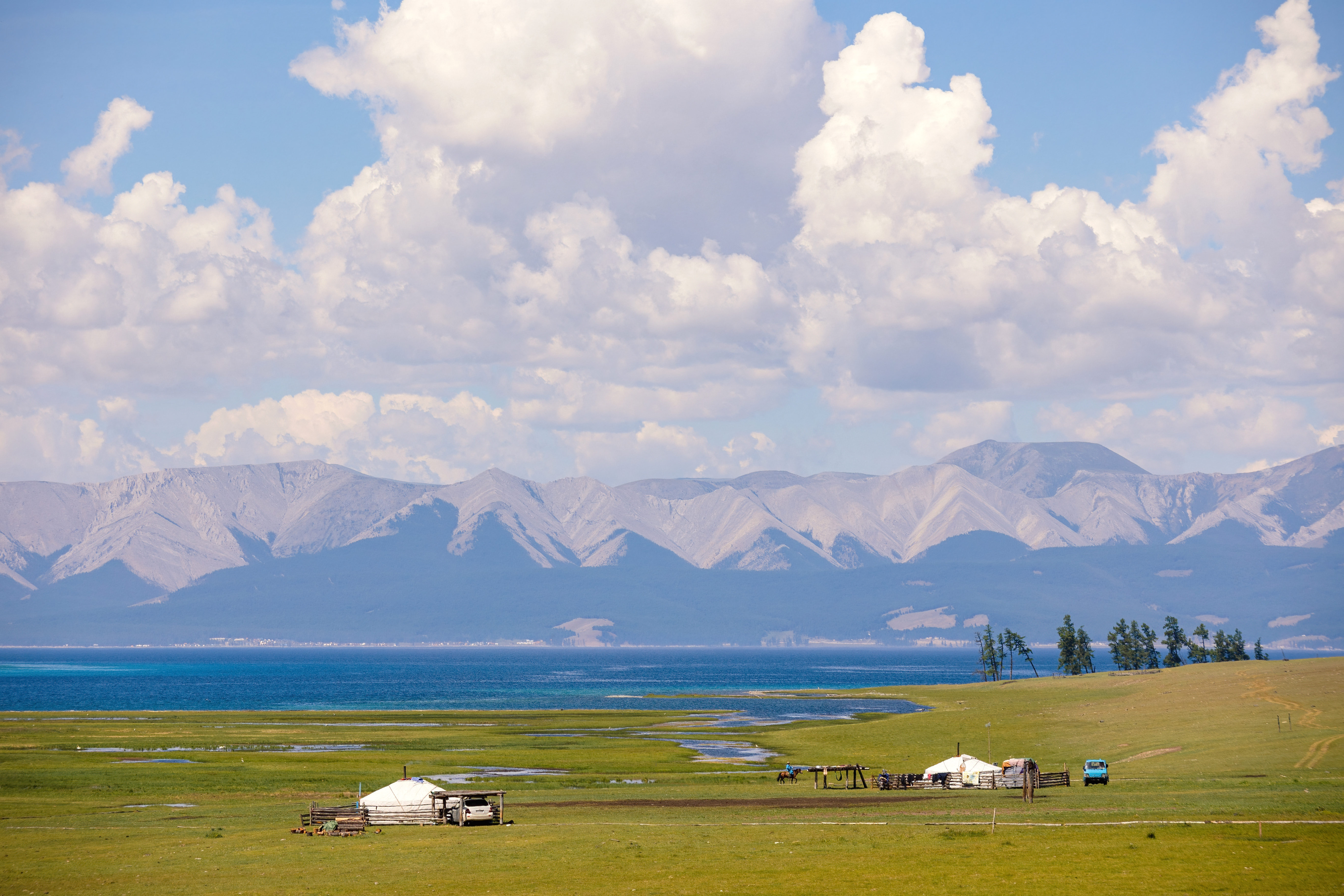 Yurts and vehicles on a grassy shore of Khövsgöl Lake, Mongolia, with turquoise water and a mountain range behind