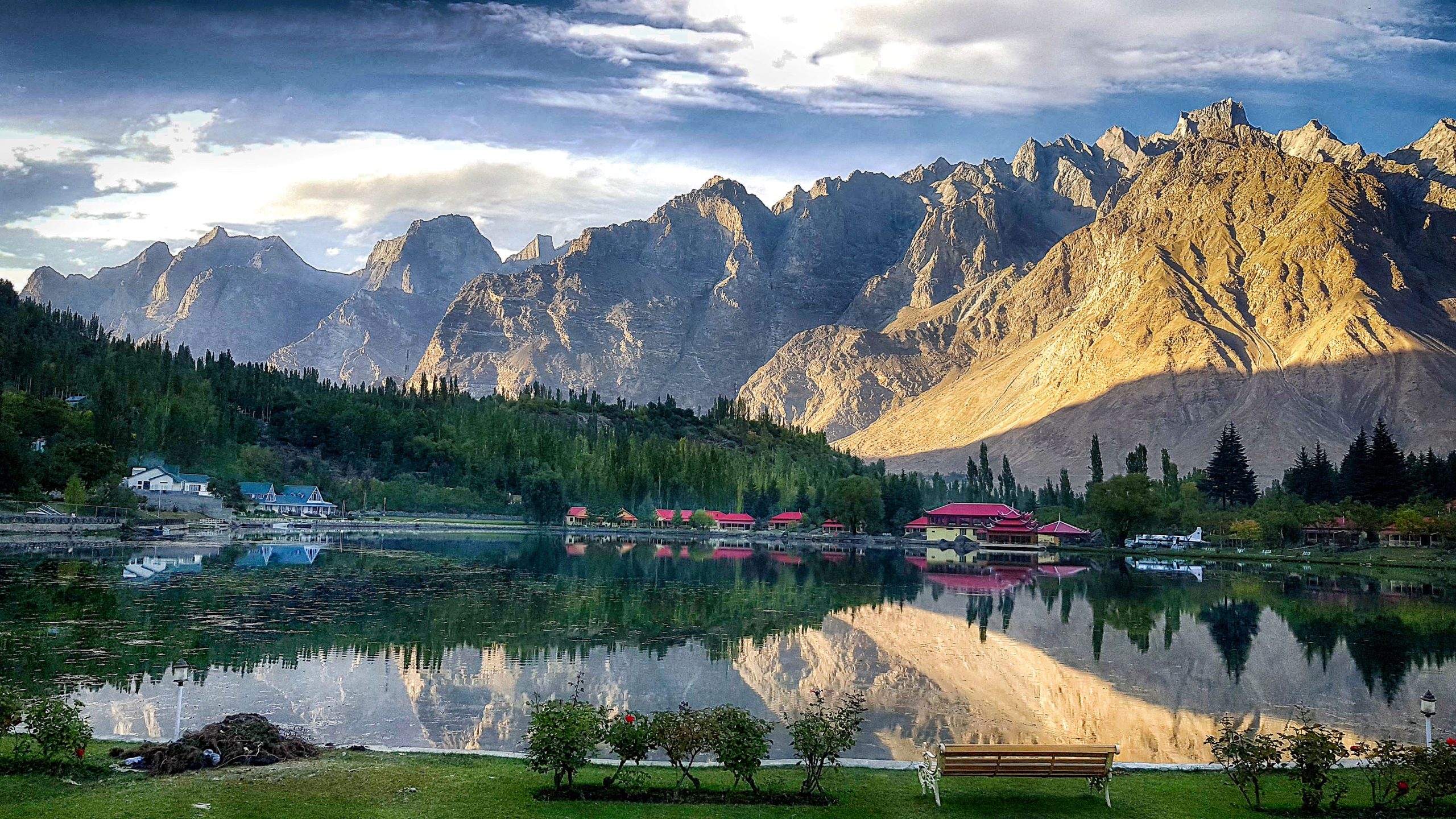 Dramatic Karakoram peaks reflected in calm lake waters at golden hour with resort buildings featuring red roofs along the shore