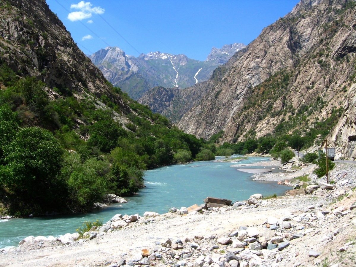 Turquoise glacial river cutting through a steep, rocky mountain valley in the Varzob Valley, with snow-streaked peaks in the distance
