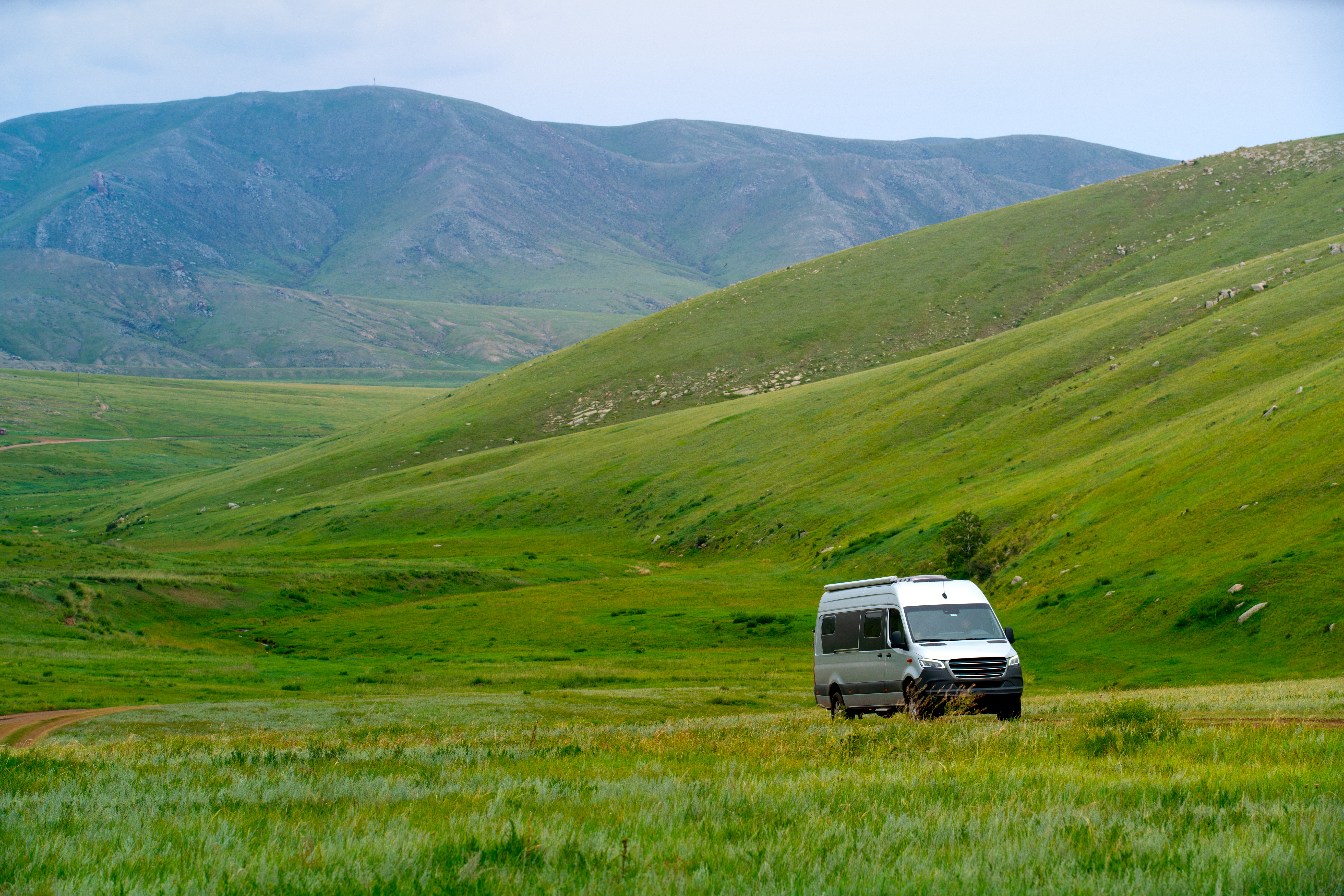 A white camper van parked on green grass at the base of grassy ridges and birch-dotted hills in Hustai National Park, Mongolia