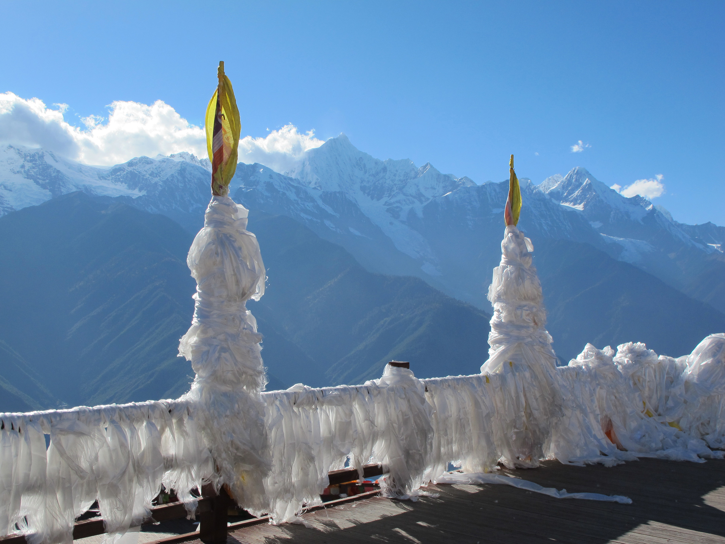 White prayer flags wrapped around poles on a viewing deck, with the snow-capped peaks of Meili Snow Mountain rising behind in Yunnan, China