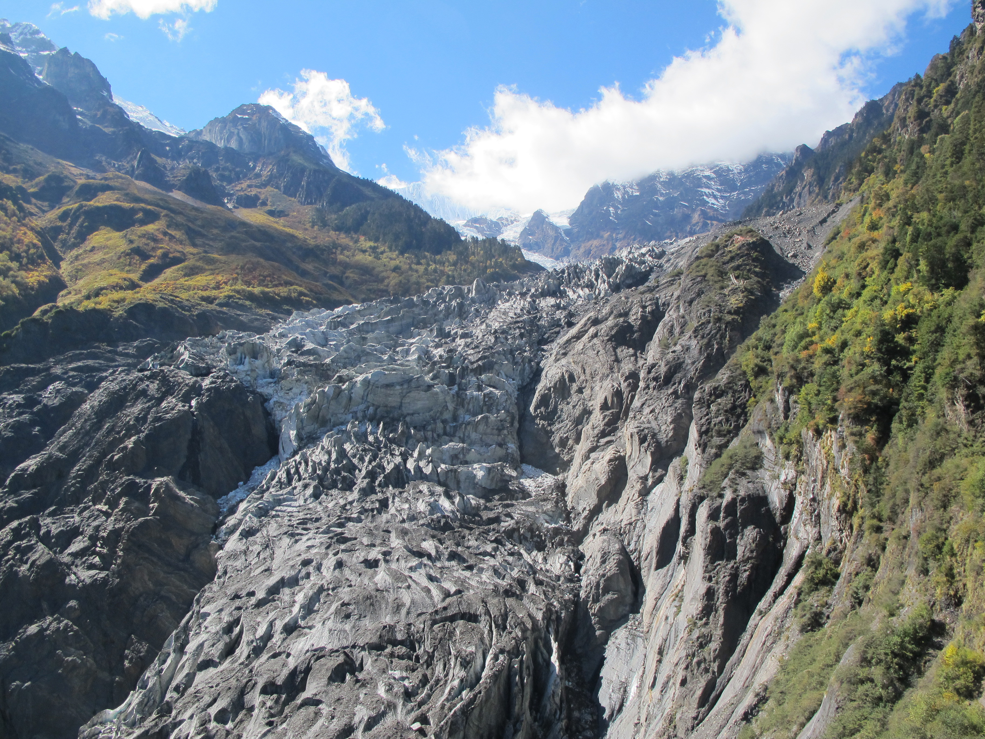 Crevassed grey ice of the Mingyong Glacier descending through a steep forested valley below snow-dusted peaks in Yunnan, China