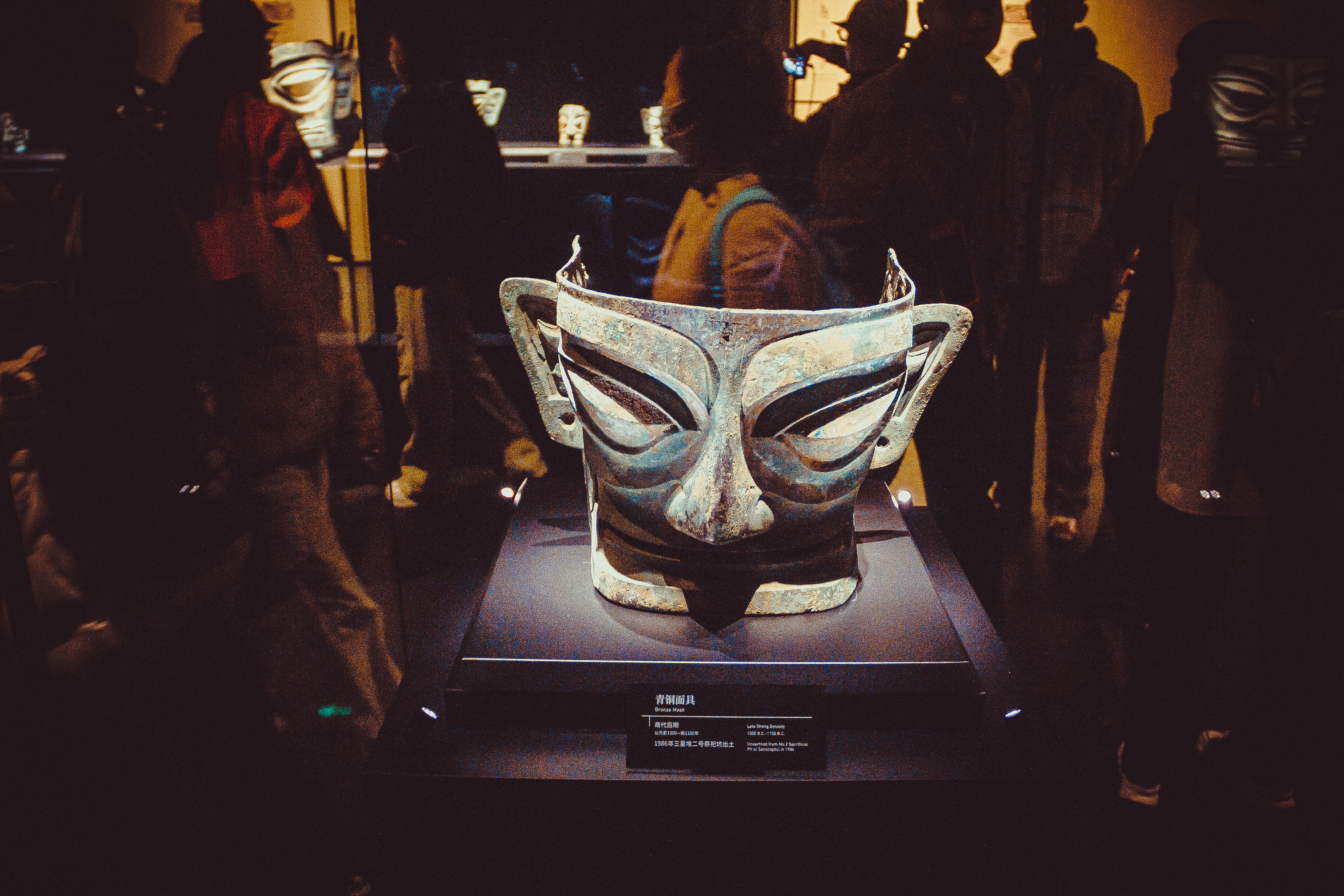 Bronze mask with protruding eyes and ears from Sanxingdui, displayed on a lit museum plinth as visitors gather around