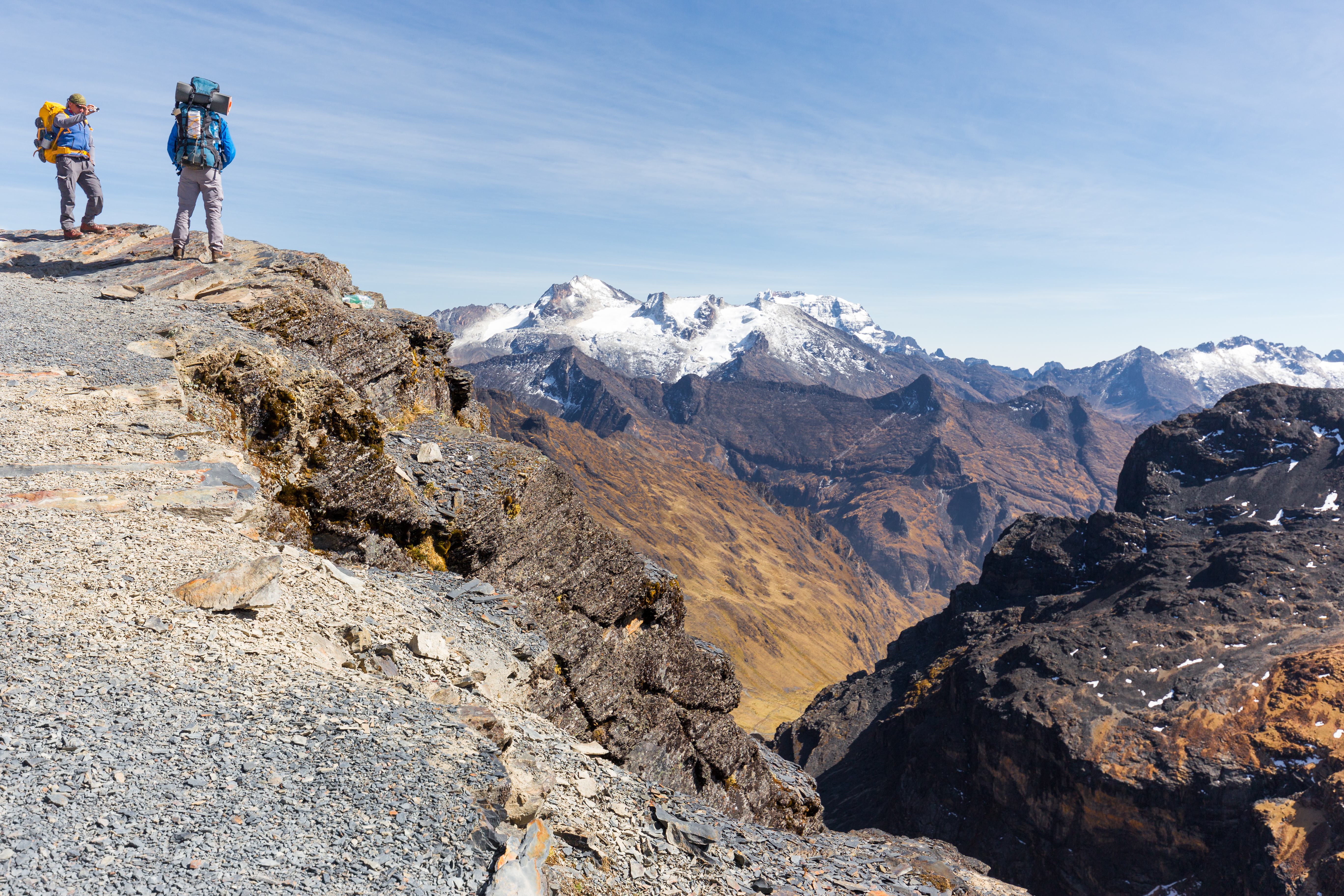 Two backpackers stand at a rocky ridge edge on the El Choro trek, overlooking snow-capped Cordillera Real peaks and a deep valley in Bolivia