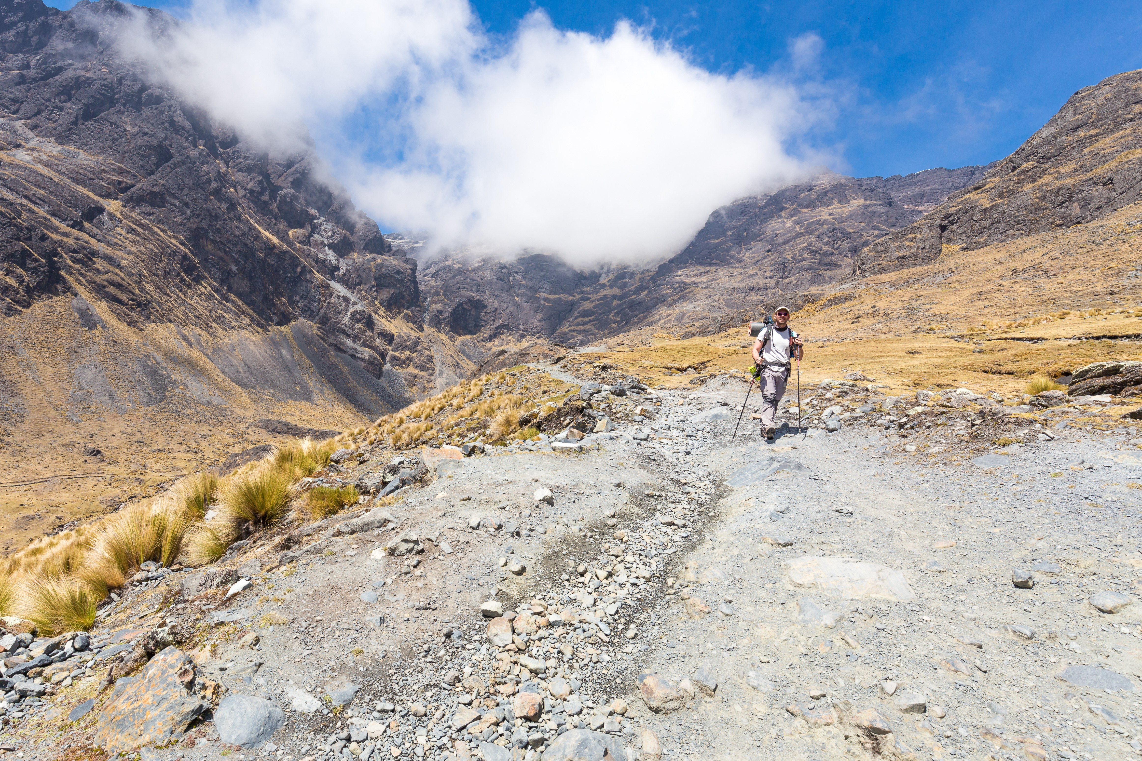 Hiker with trekking poles and backpack walks a rocky trail on the El Choro trek, high Andean peaks and cloud behind