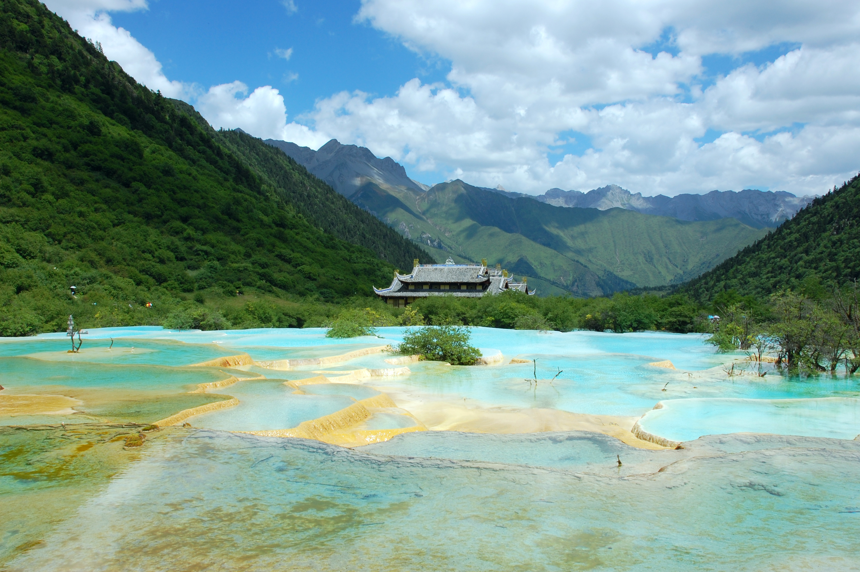 Blue and gold travertine terrace pools at Huanglong with a Ming-dynasty temple roof and forested peaks behind