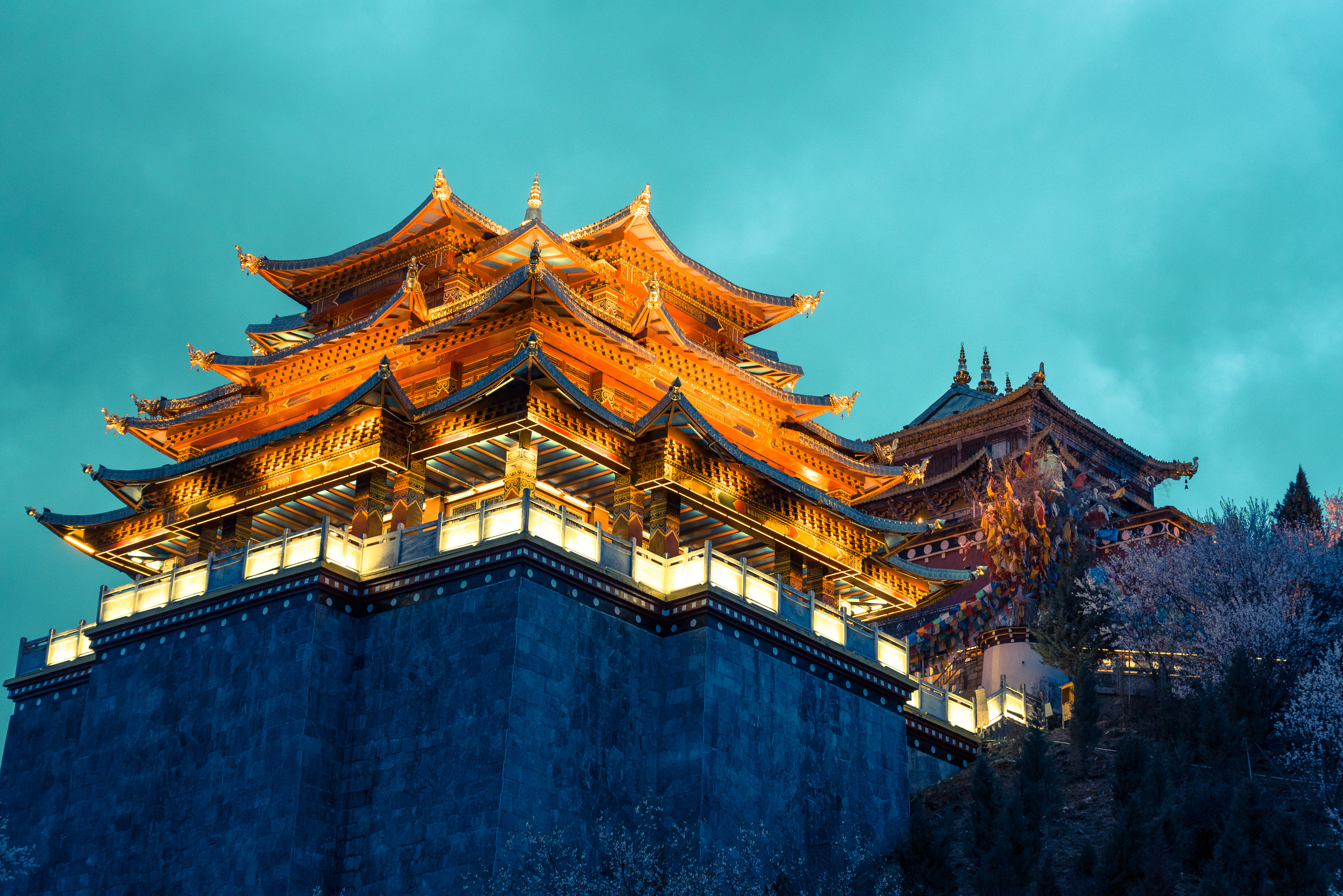 Gilded, multi-tiered roofs of Songzanlin Monastery illuminated at night against a teal evening sky, stone walls below