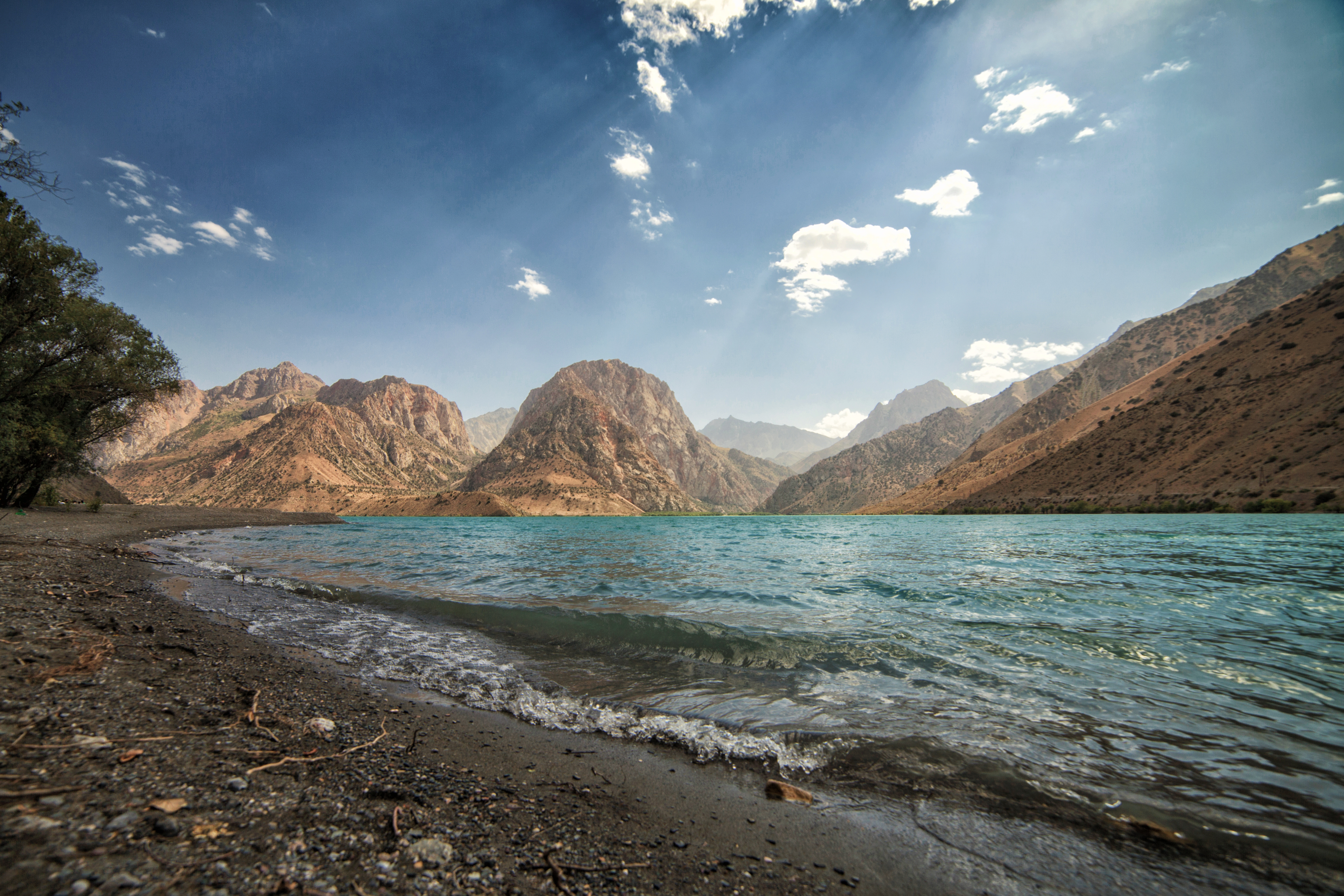 Turquoise waters of Iskanderkul lake meet a pebbled shore, with sunlit peaks of the Fann Mountains rising behind under a blue sky