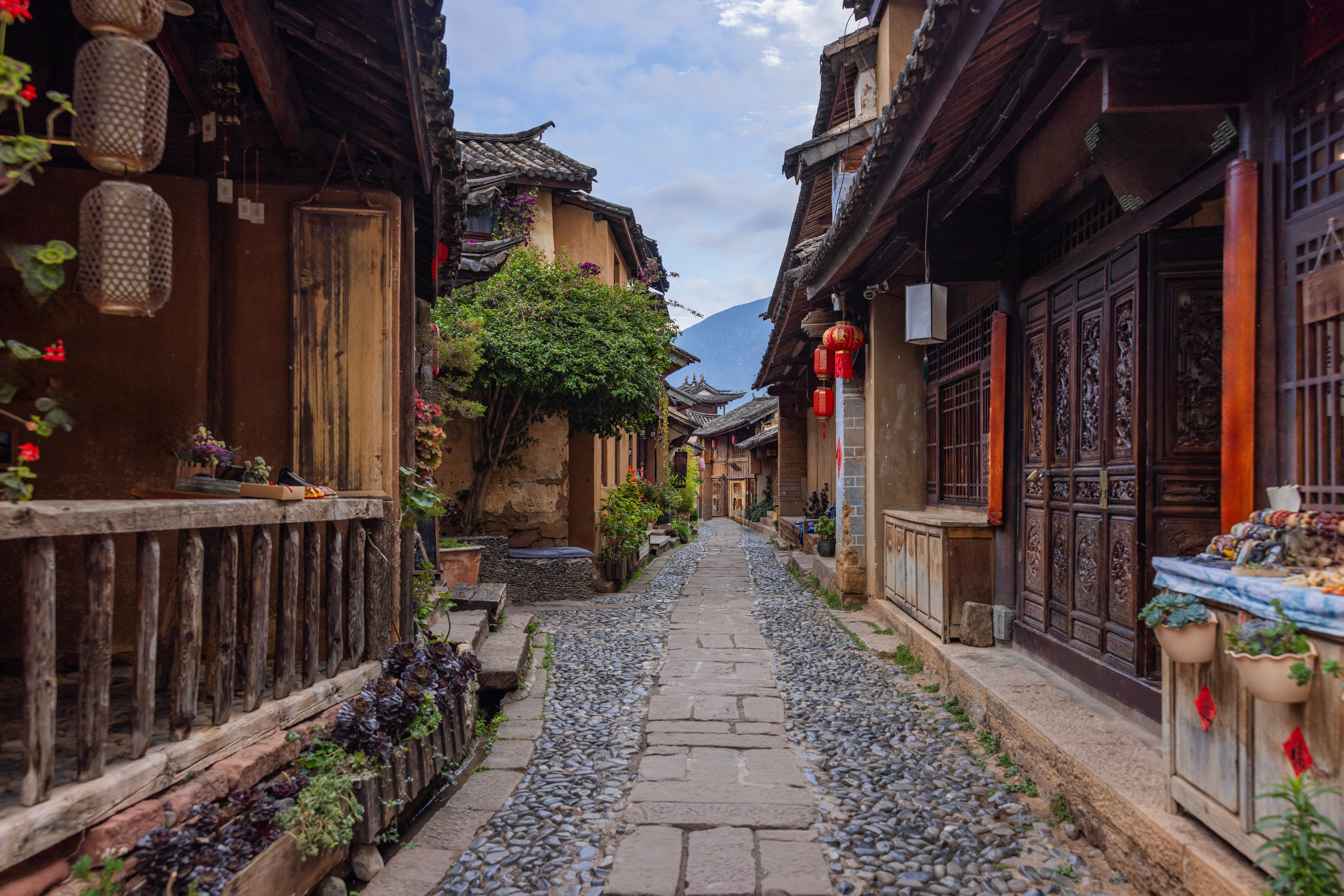 Cobblestone lane through Shaxi lined with timber houses, carved wooden doors, red lanterns and potted plants, mountains visible ahead