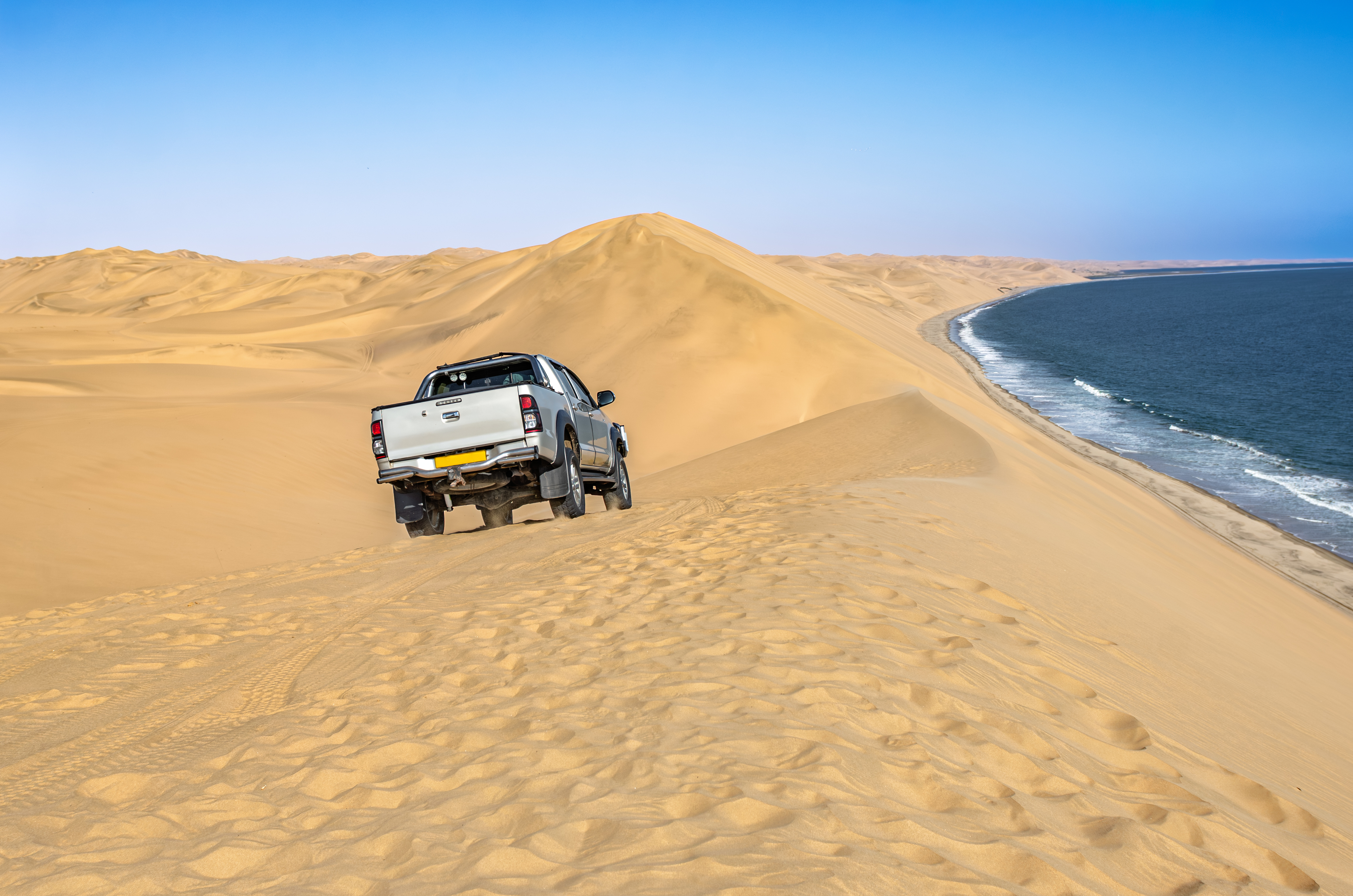 A 4x4 vehicle crests a sand dune at Sandwich Harbour, with the Atlantic coastline curving away in the background