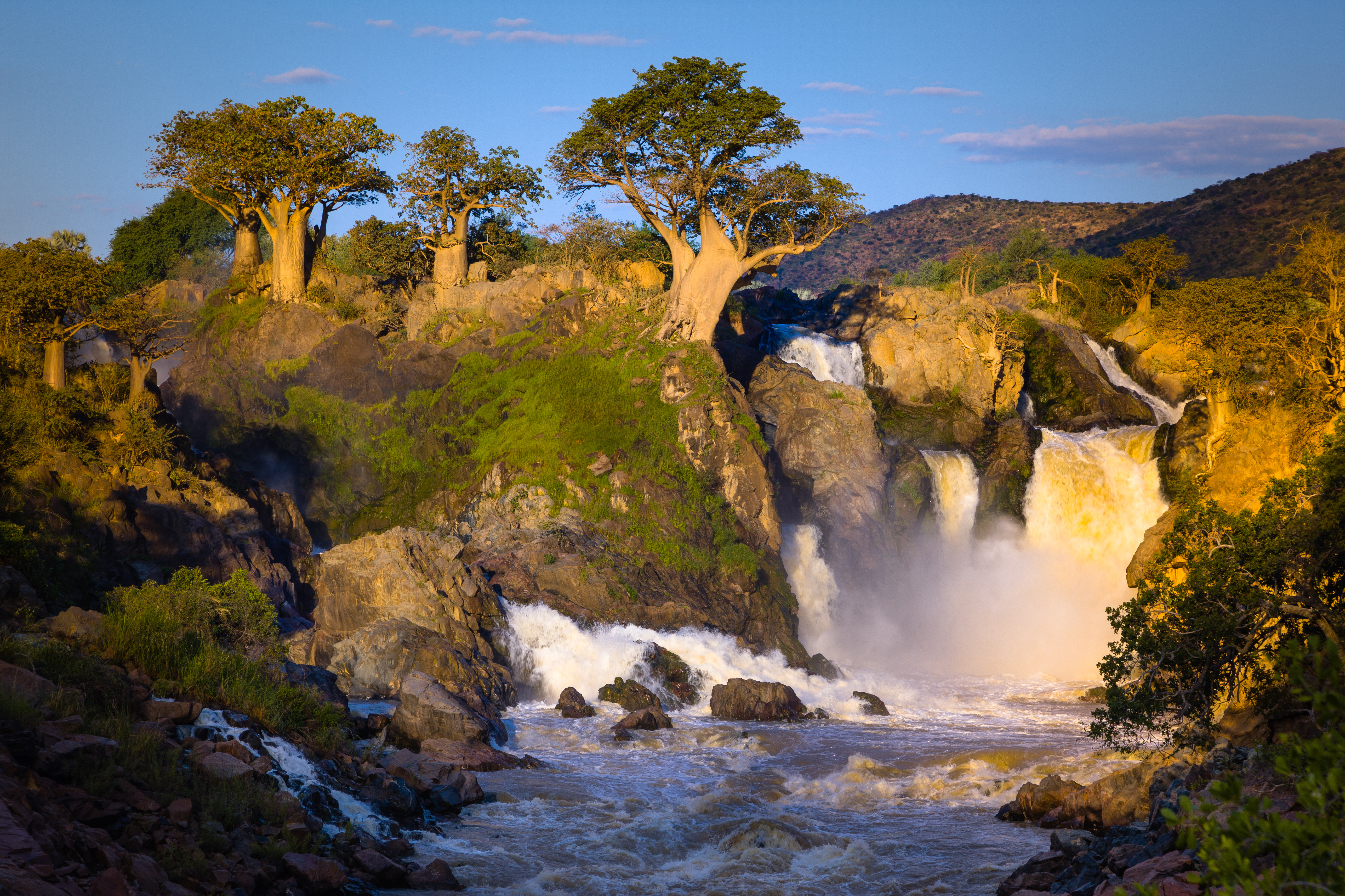 Waterfall cascading over dark rock at Epupa Falls, with baobab trees rising from the rocky outcrop above the falling water