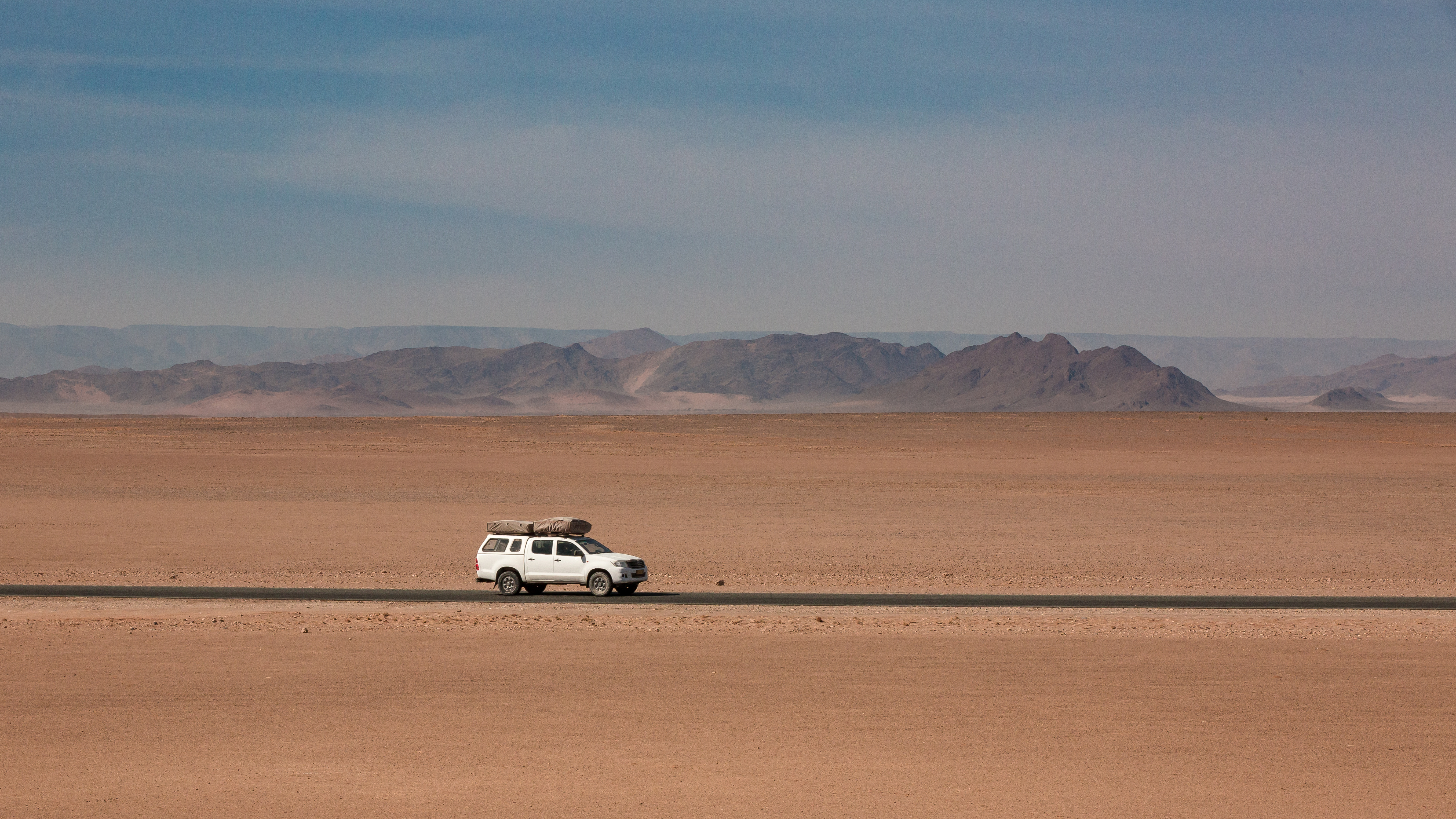 A white pickup truck loaded with gear drives a paved road through the open gravel plains of the Namib Desert, distant mountains behind