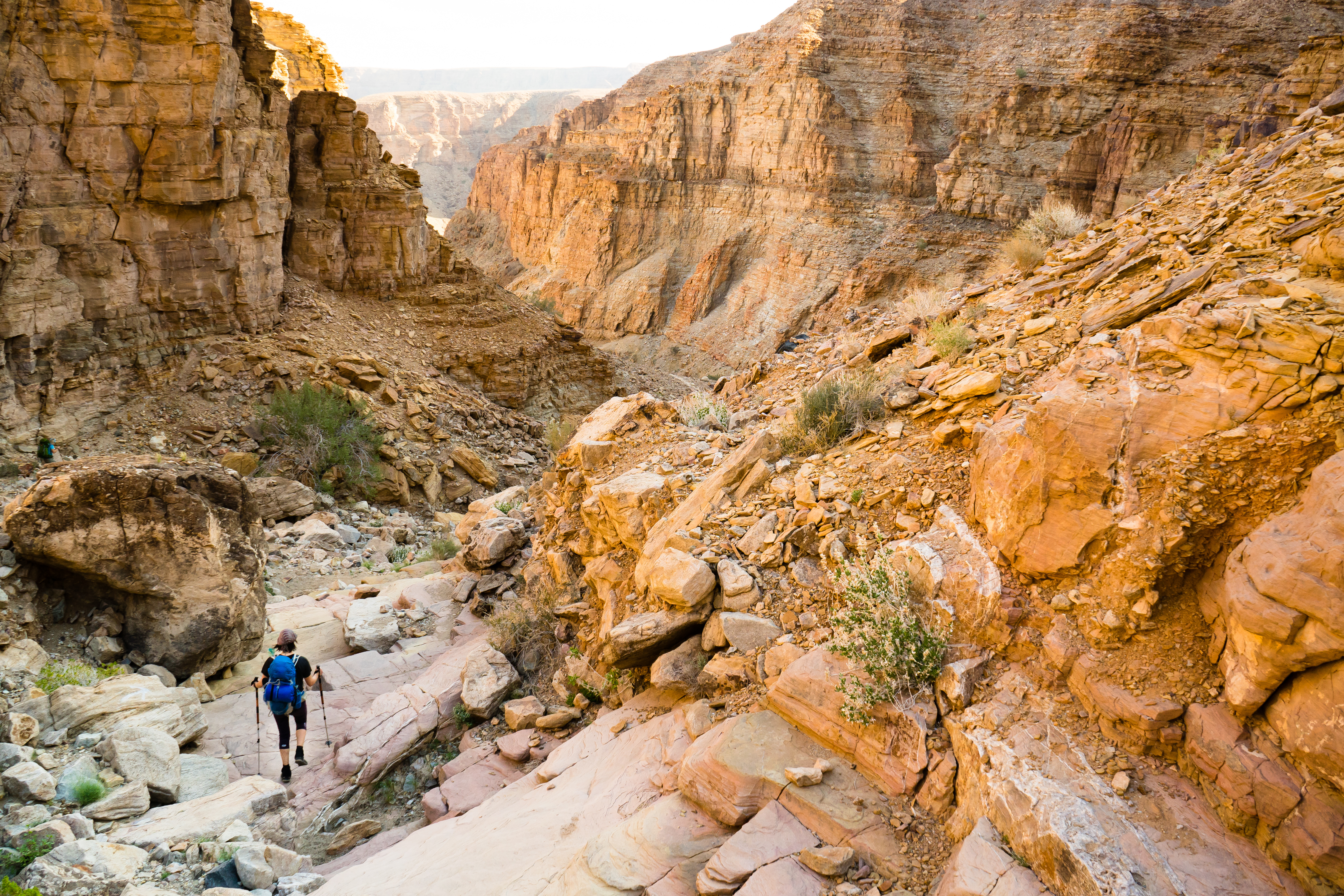 Hiker with trekking poles descends a rocky trail into the orange-walled depths of Fish River Canyon in Namibia