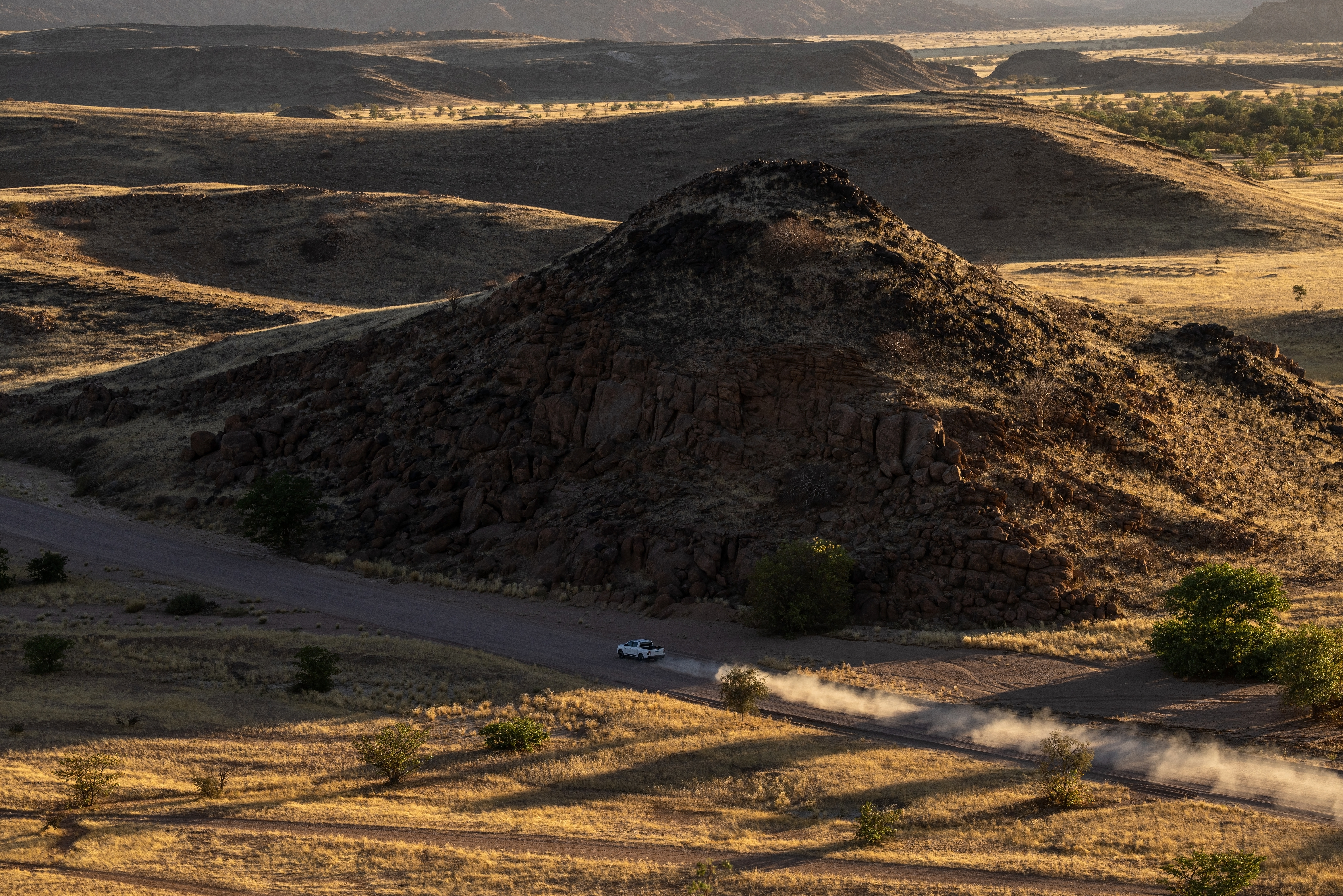 A vehicle kicks up dust on a dirt road winding past a rocky hill in Damaraland, Namibia, seen from above