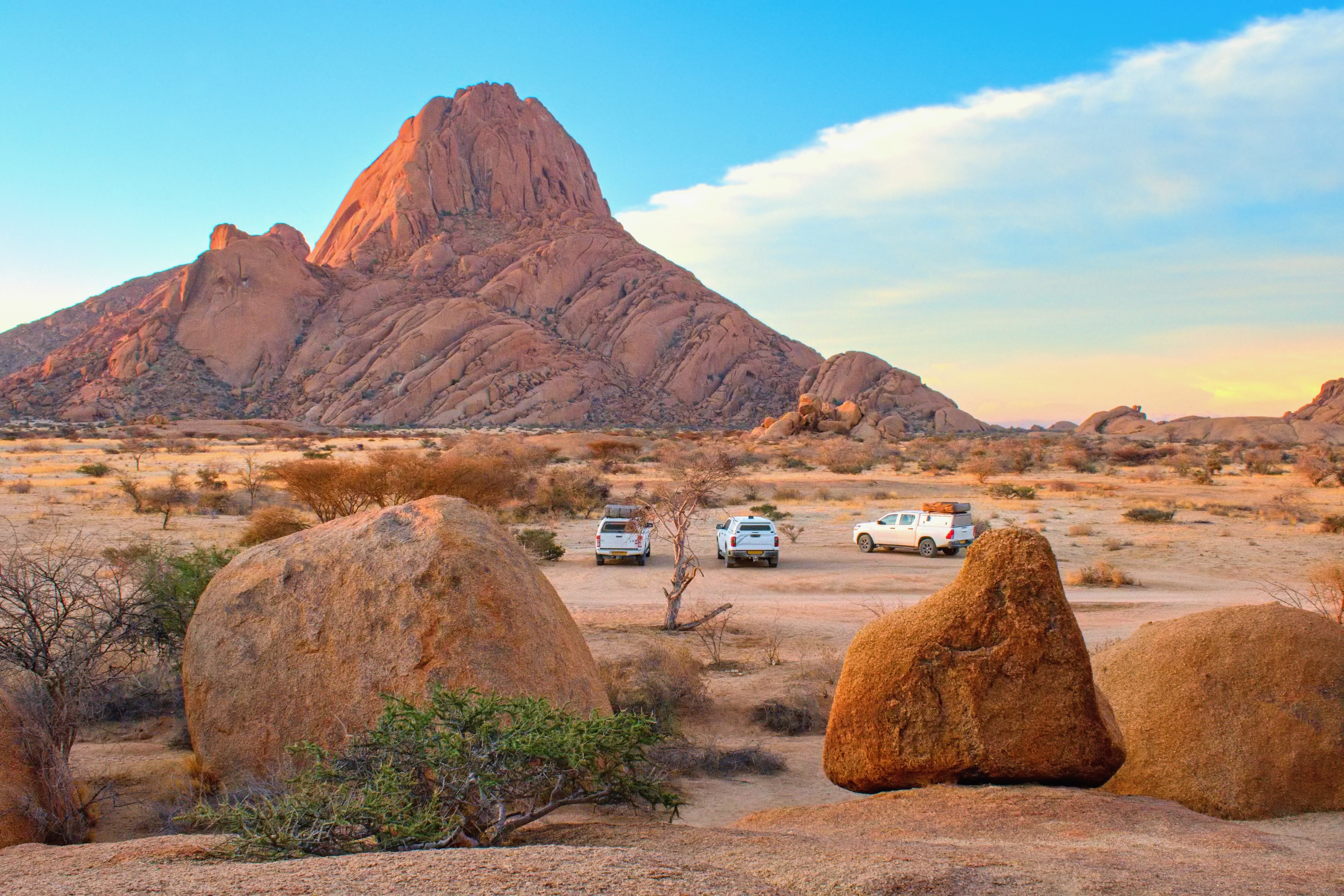 Granite peak of Spitzkoppe rises over a desert plain with three parked 4x4 vehicles at a wild campsite