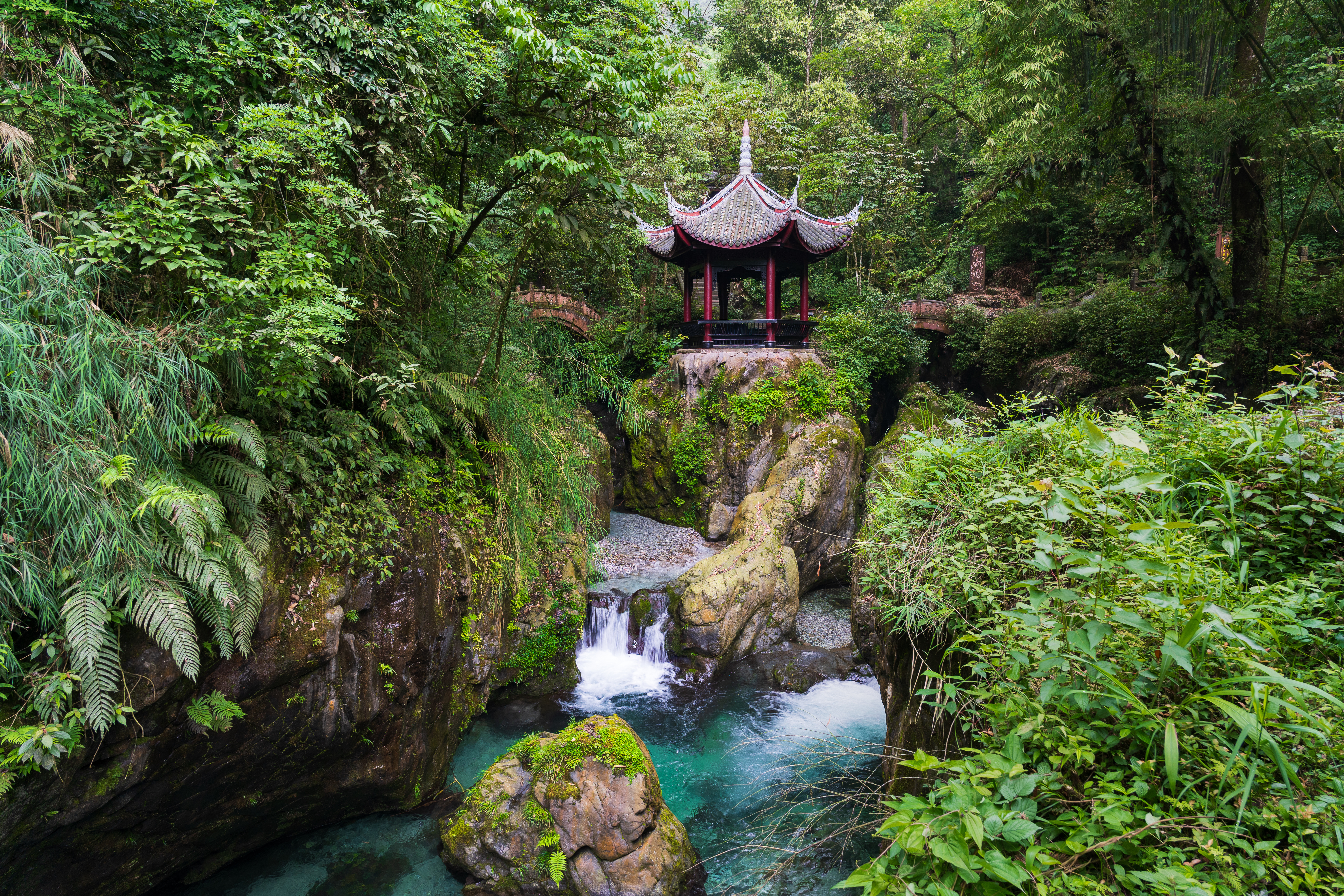 A red and grey Chinese pavilion perches on rock above a turquoise stream and small waterfall, surrounded by dense forest on Mount Emei