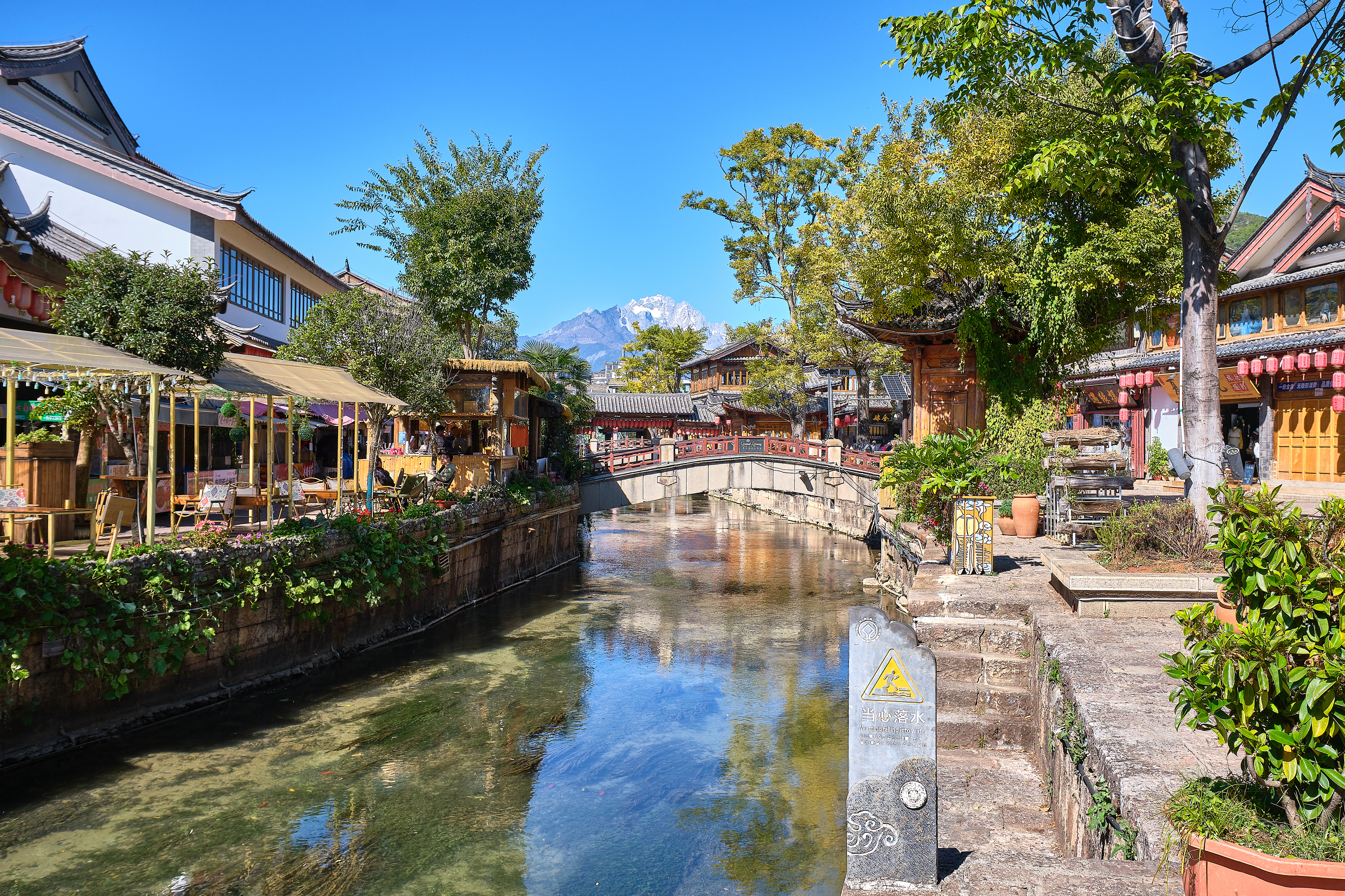 Canal lined with cafes and wooden buildings in Lijiang old town, a stone bridge ahead and snow capped Jade Dragon Mountain on the horizon