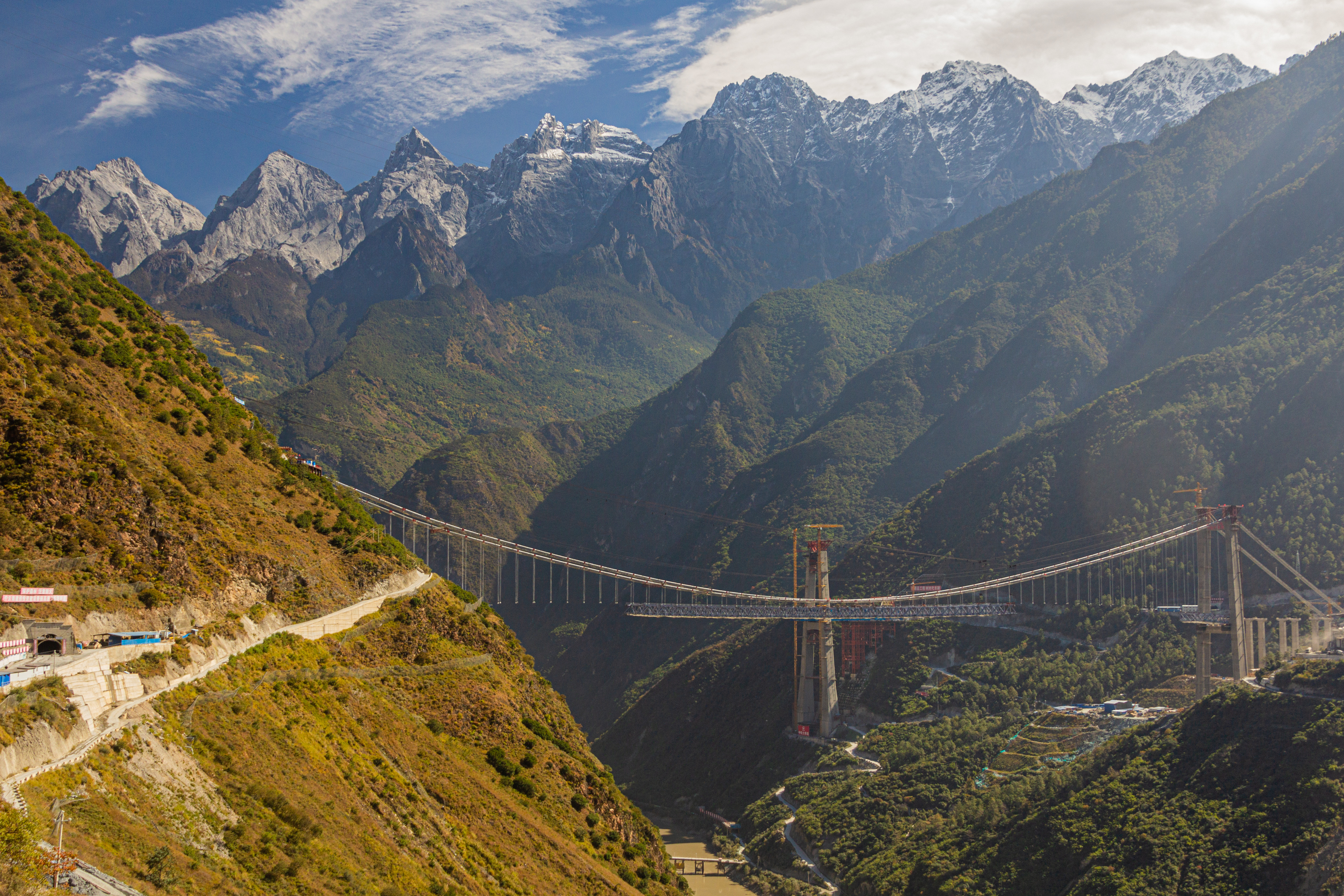 A large suspension bridge under construction spans a deep valley on the Jinsha River, with snow-capped peaks rising behind, Yunnan, China