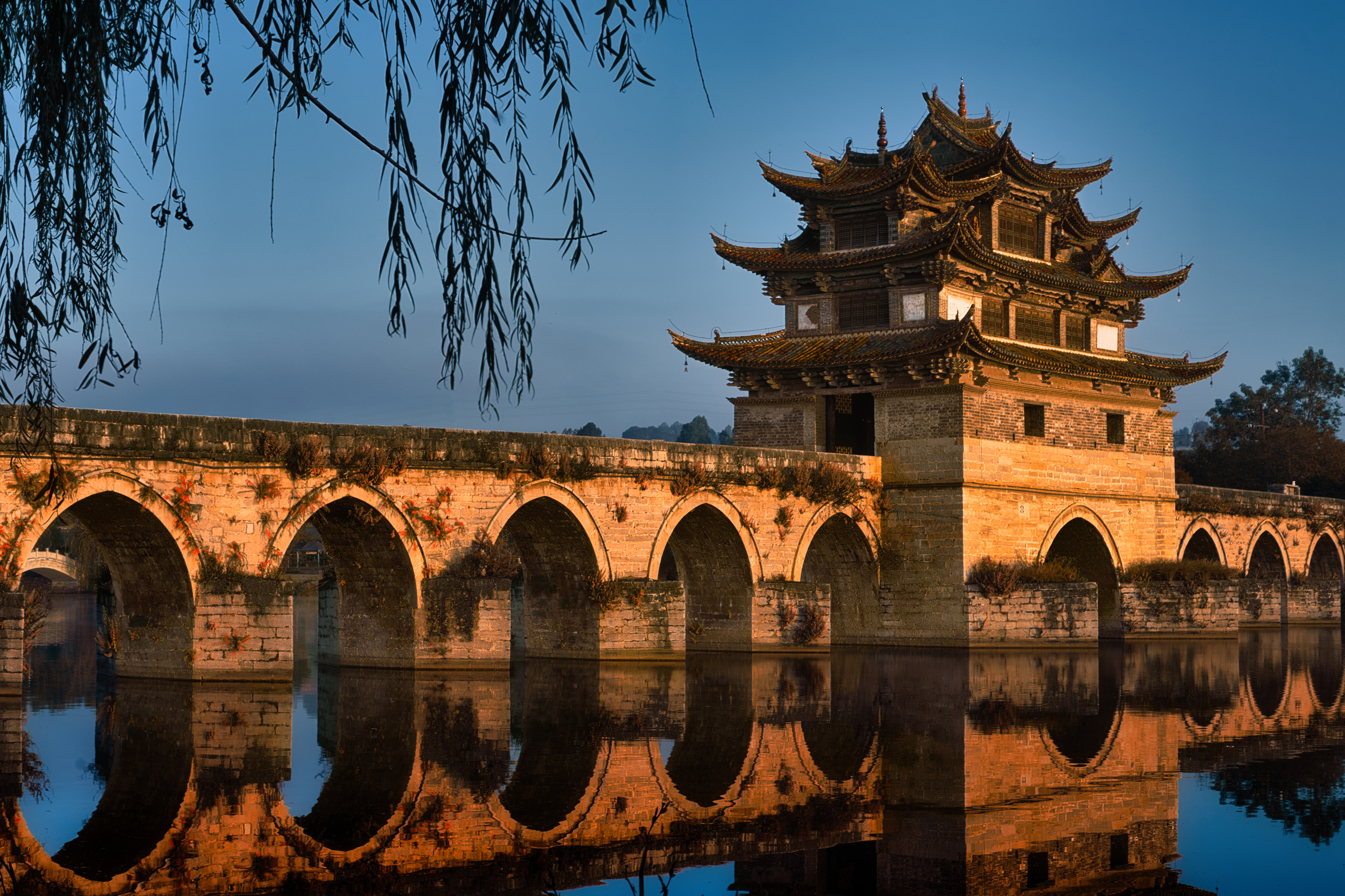 Multi-arched stone Double Dragon Bridge in Jianshui reflected in still water, its central pavilion lit gold by sunset light