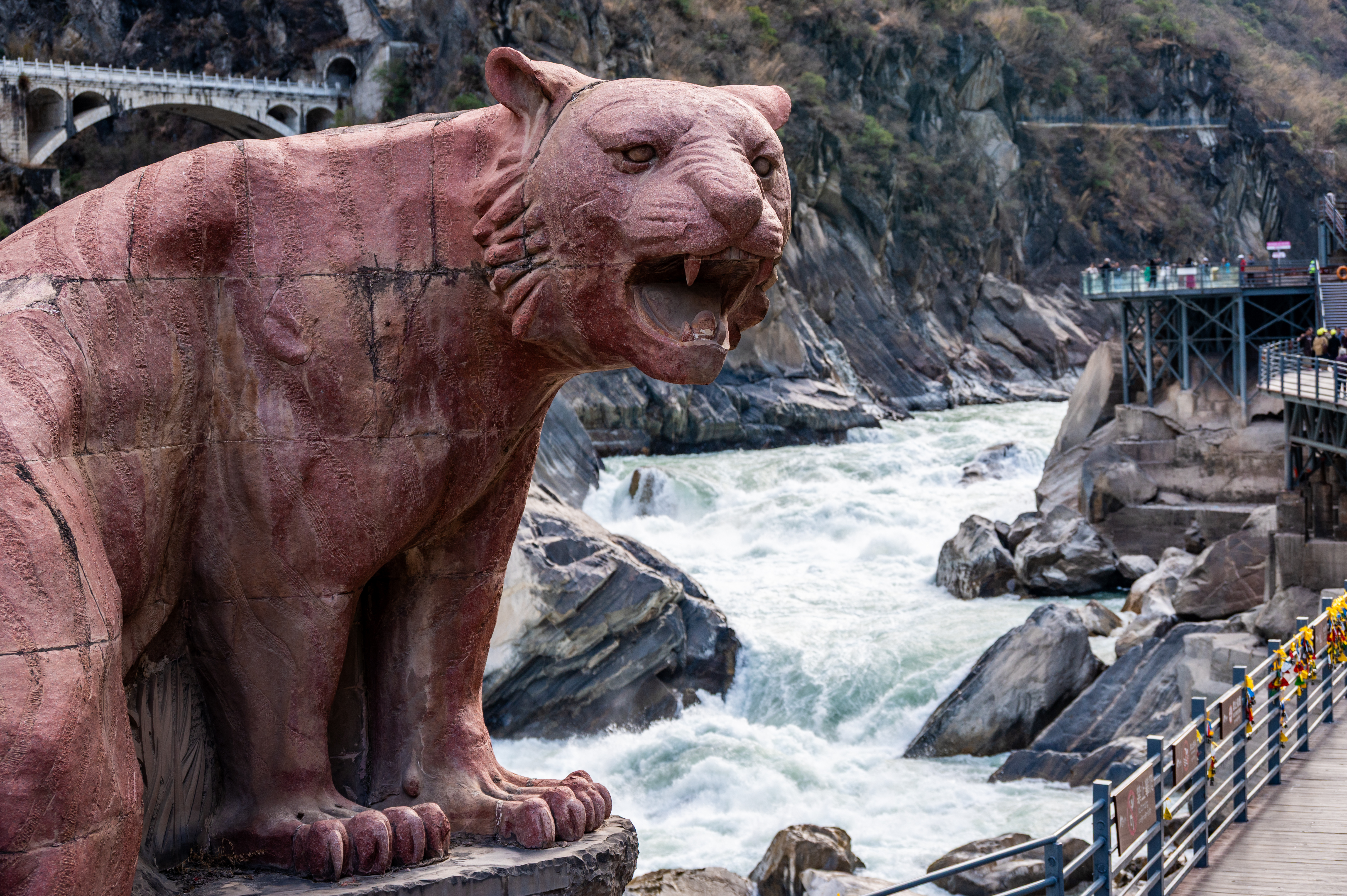 Carved stone tiger statue perched above the rushing rapids of Tiger Leaping Gorge, with a viewing walkway and bridge in the background