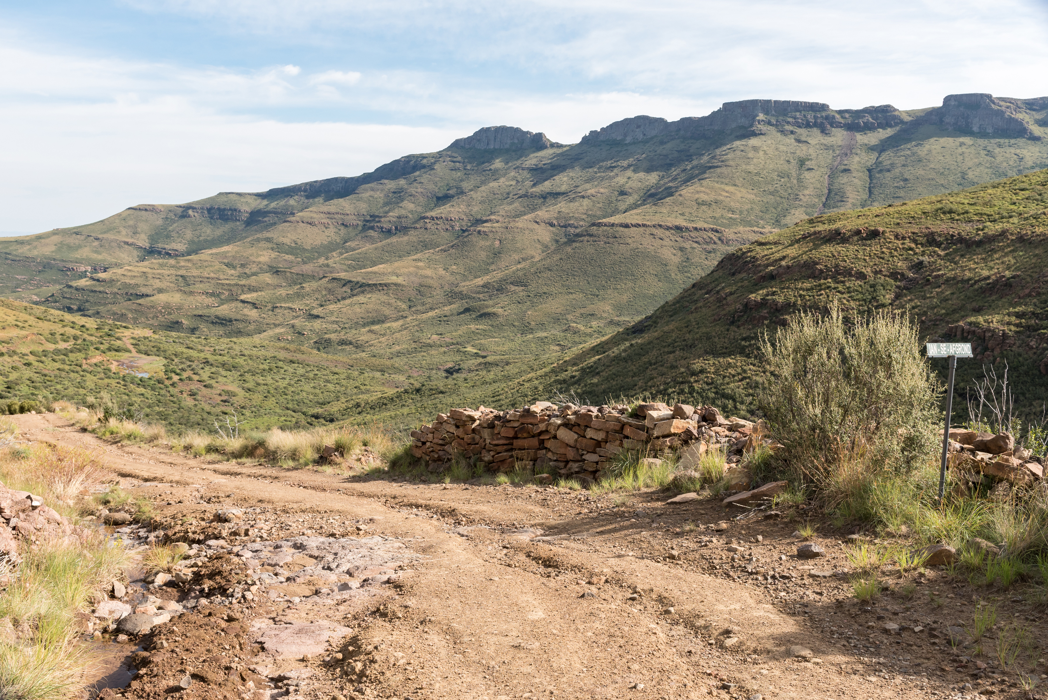 Rutted dirt track winding through a mountain pass in Namibia, with a stone cairn marker and layered escarpments rising in the distance