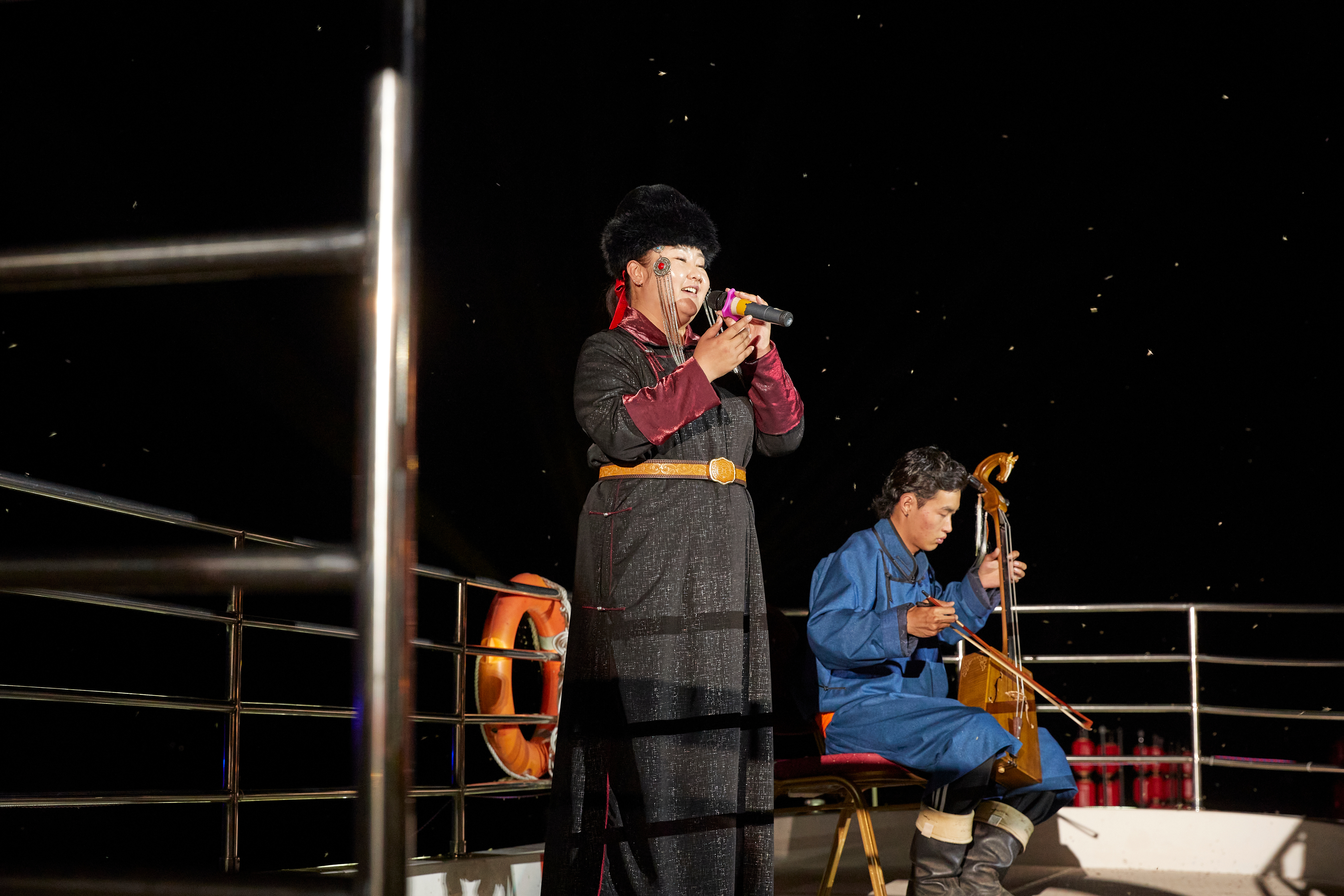 Singer in traditional Mongolian dress with a microphone performs beside a morin khuur player at night under a starry sky