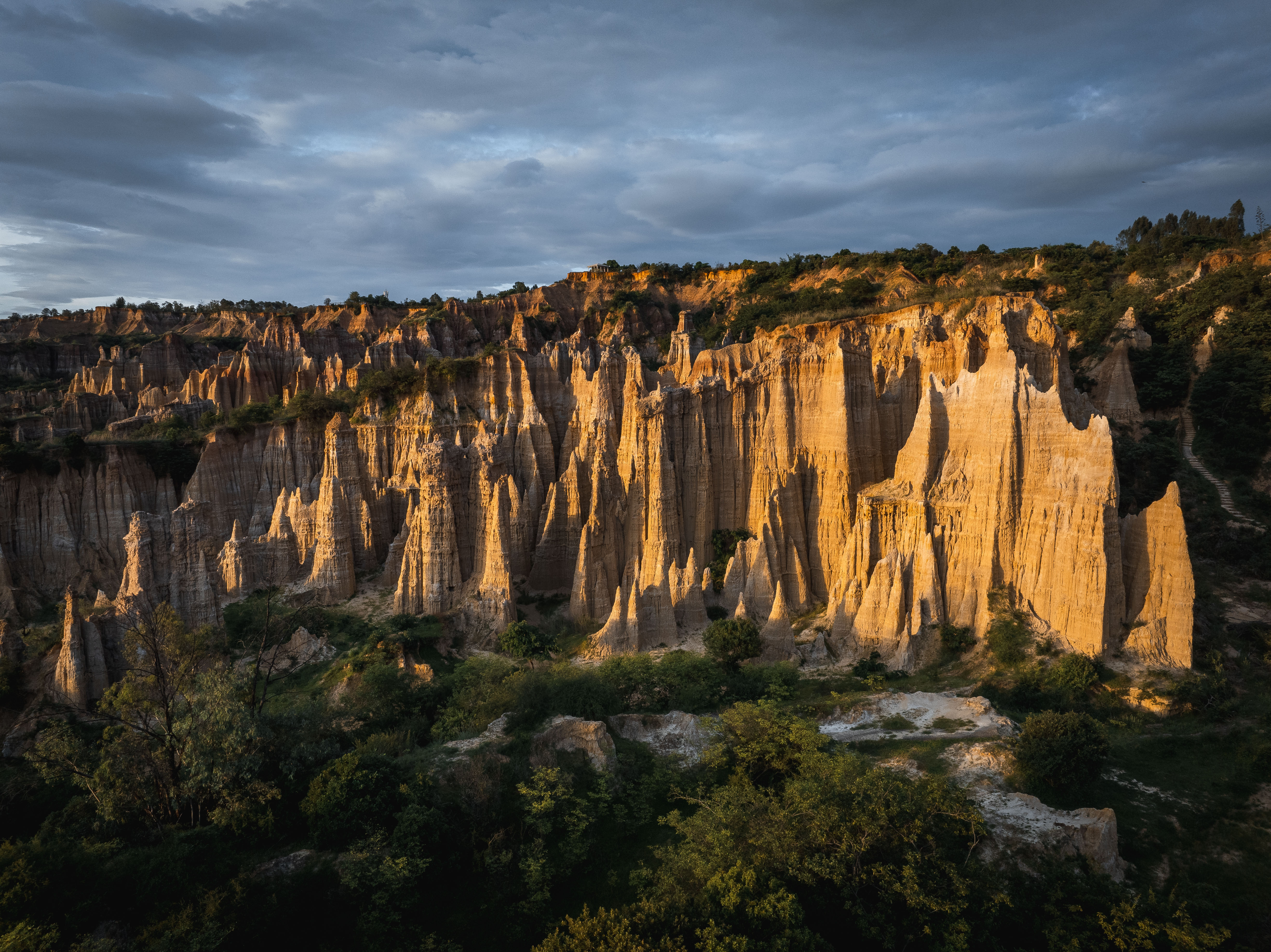Sunset light rakes tall eroded earth pinnacles at the Yuanmou Earth Forest in Yunnan, with green scrub at their base beneath a cloudy sky