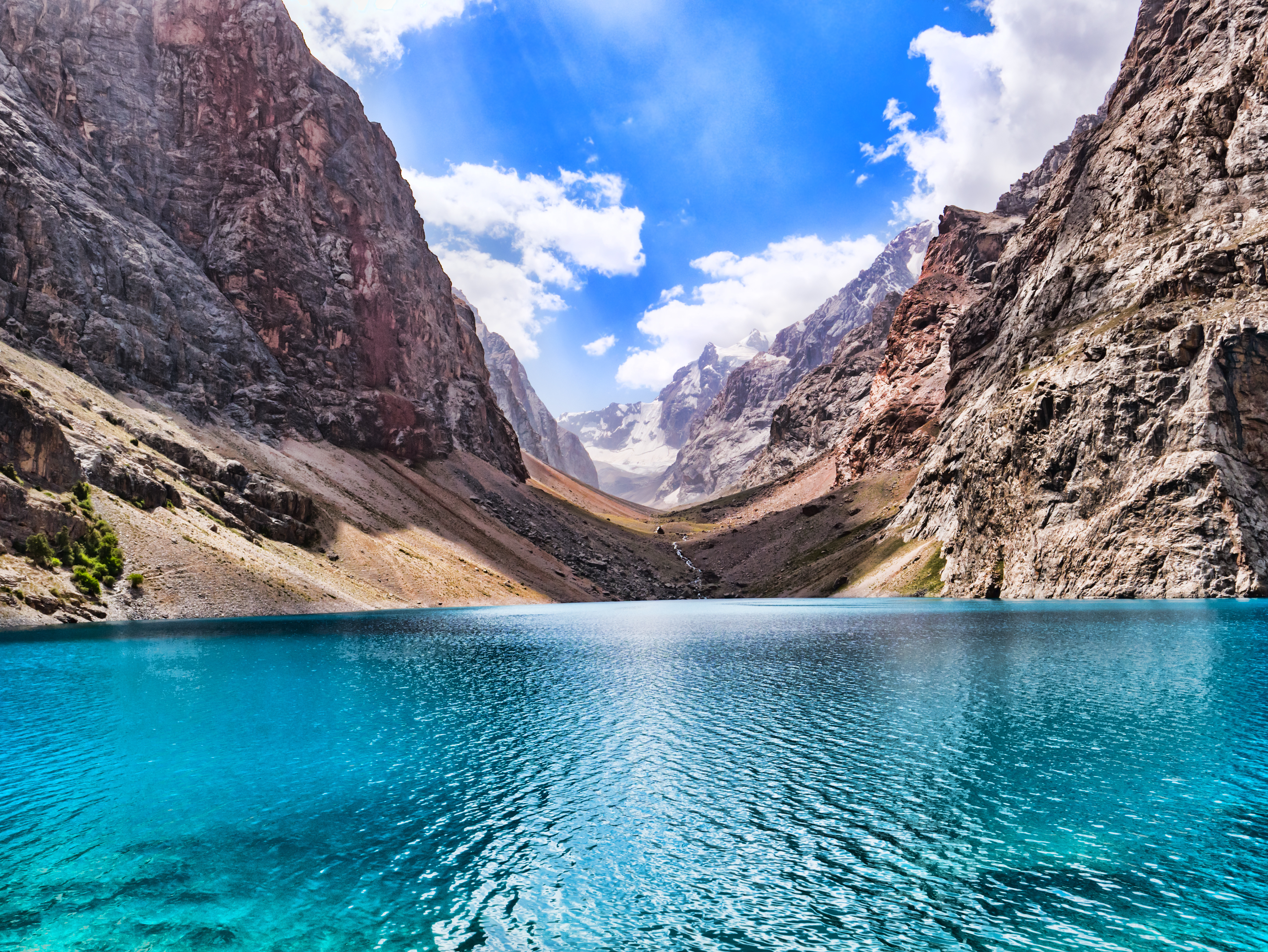 Turquoise waters of Big Alo Lake fill a valley floor, framed by steep rocky mountainsides and snow-capped peaks in the Fann Mountains