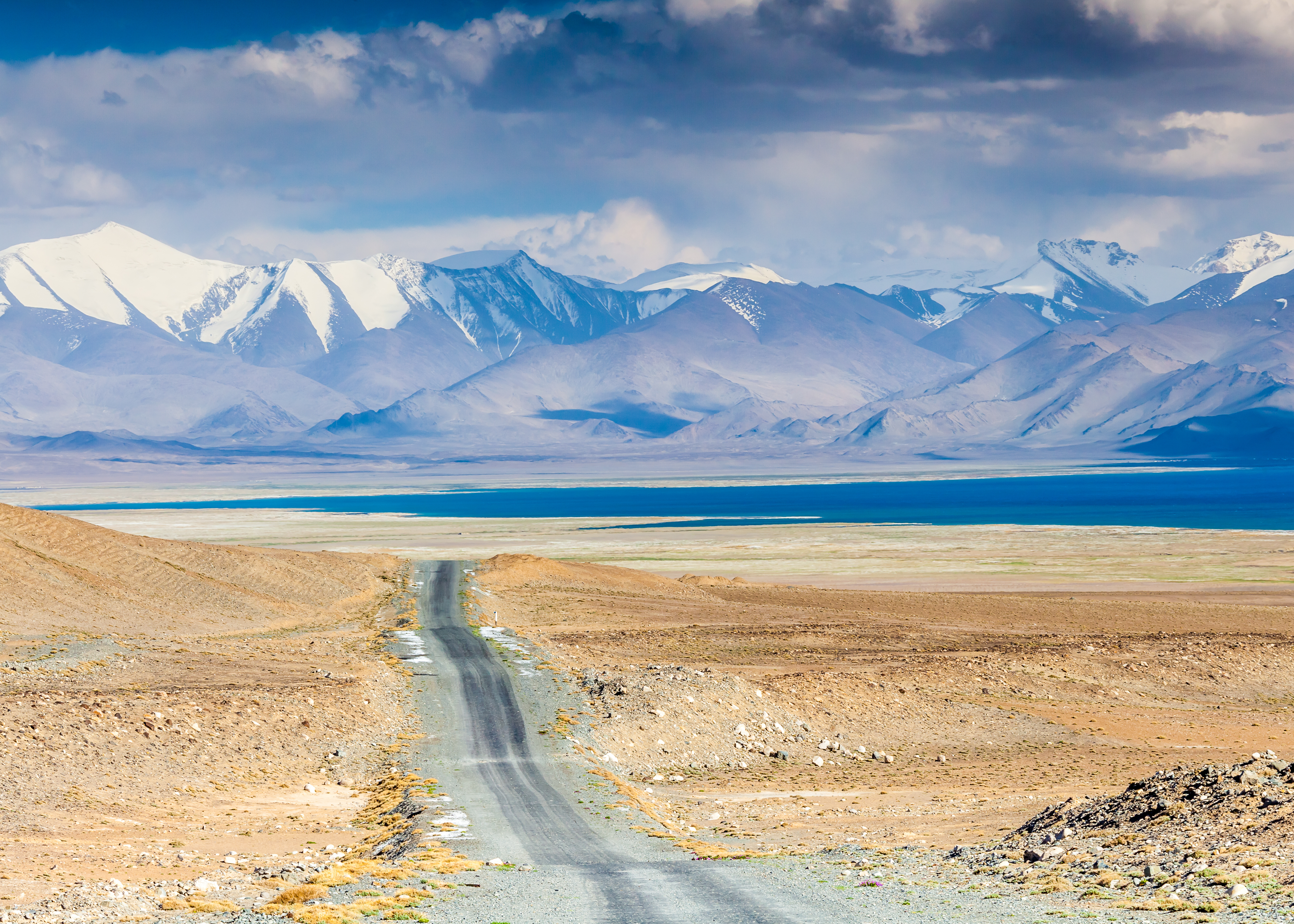 Gravel road crosses a dry plateau toward the turquoise waters of Karakul Lake, with snow-capped Pamir peaks beyond
