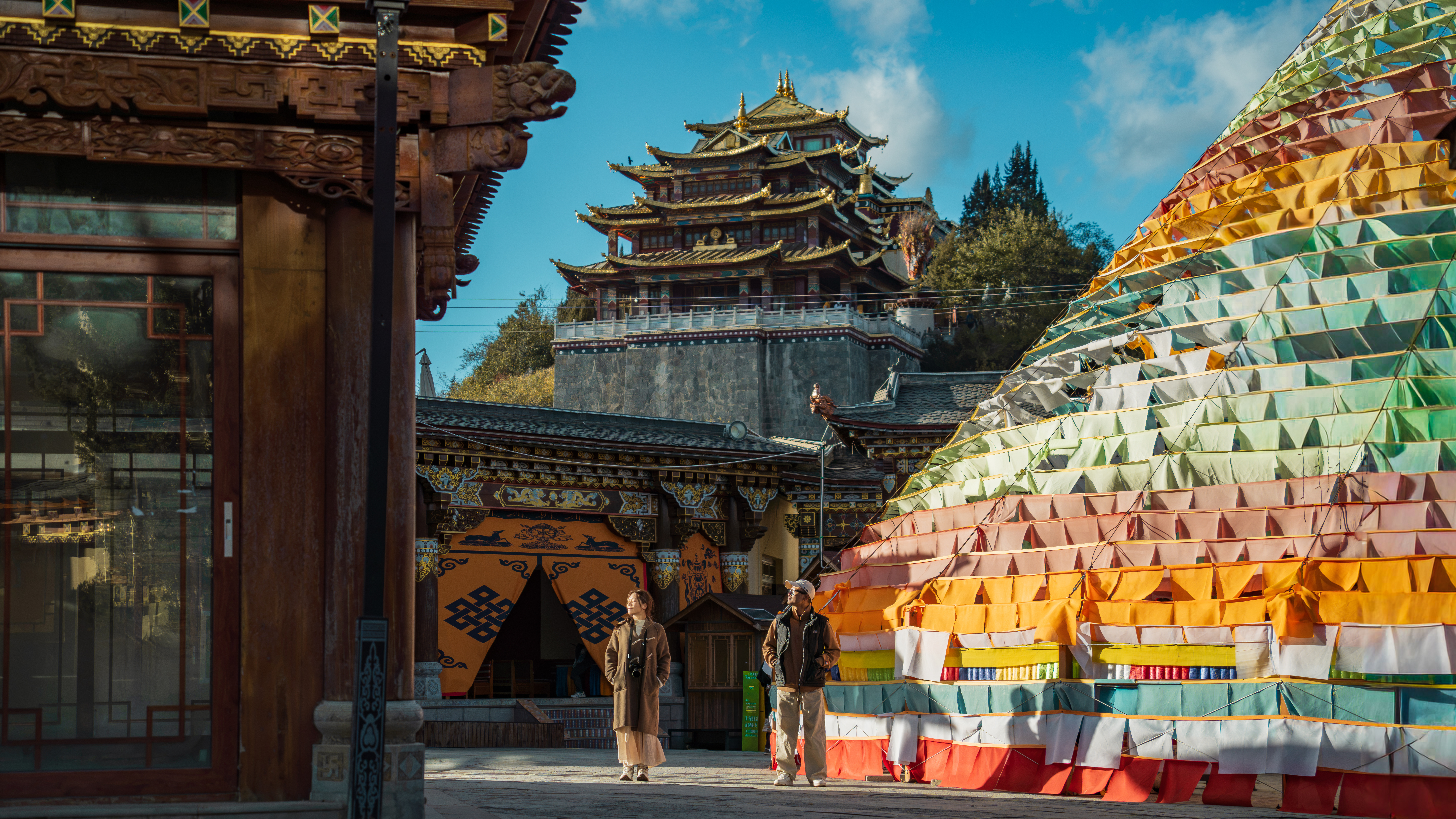 Two travelers stand before a pyramid of layered prayer flags facing a gilded, multi-tiered temple in Shangri-La Old Town, China