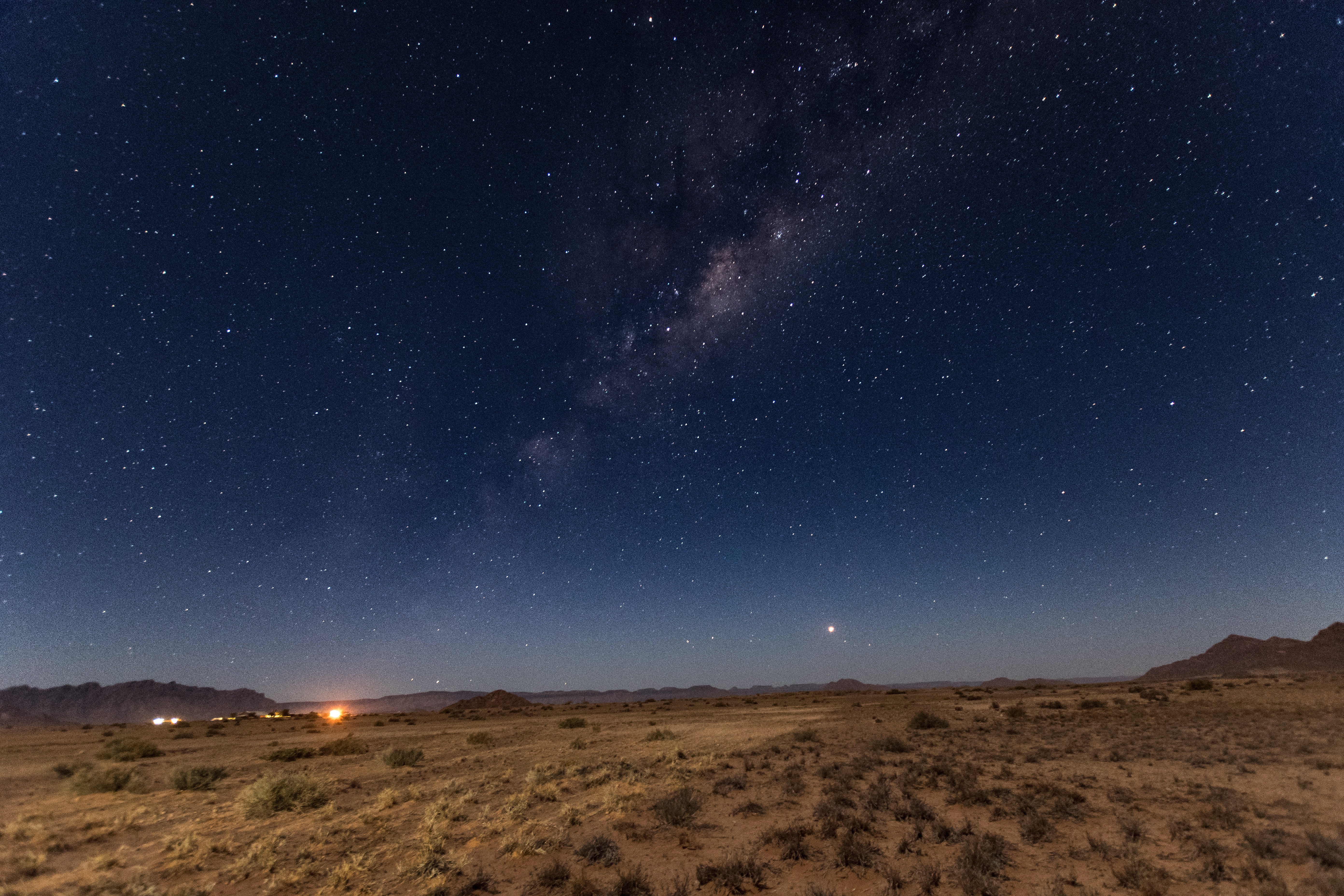 Milky Way arcs over the dark silhouette of the Namib Desert, with scattered distant lights and a bright planet low on the horizon