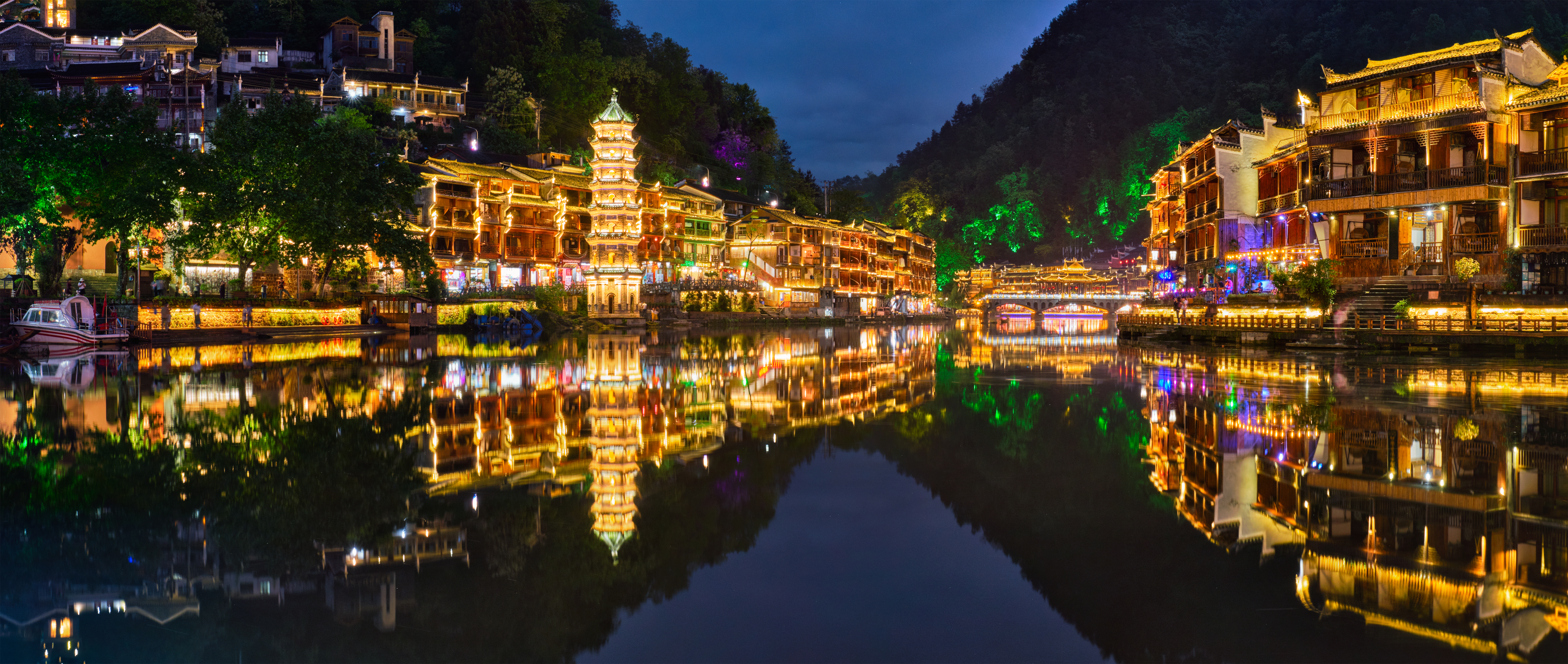 Feng Huang Ancient Town illuminated at night, riverside wooden buildings and a pagoda reflected in the still Tuo Jiang River
