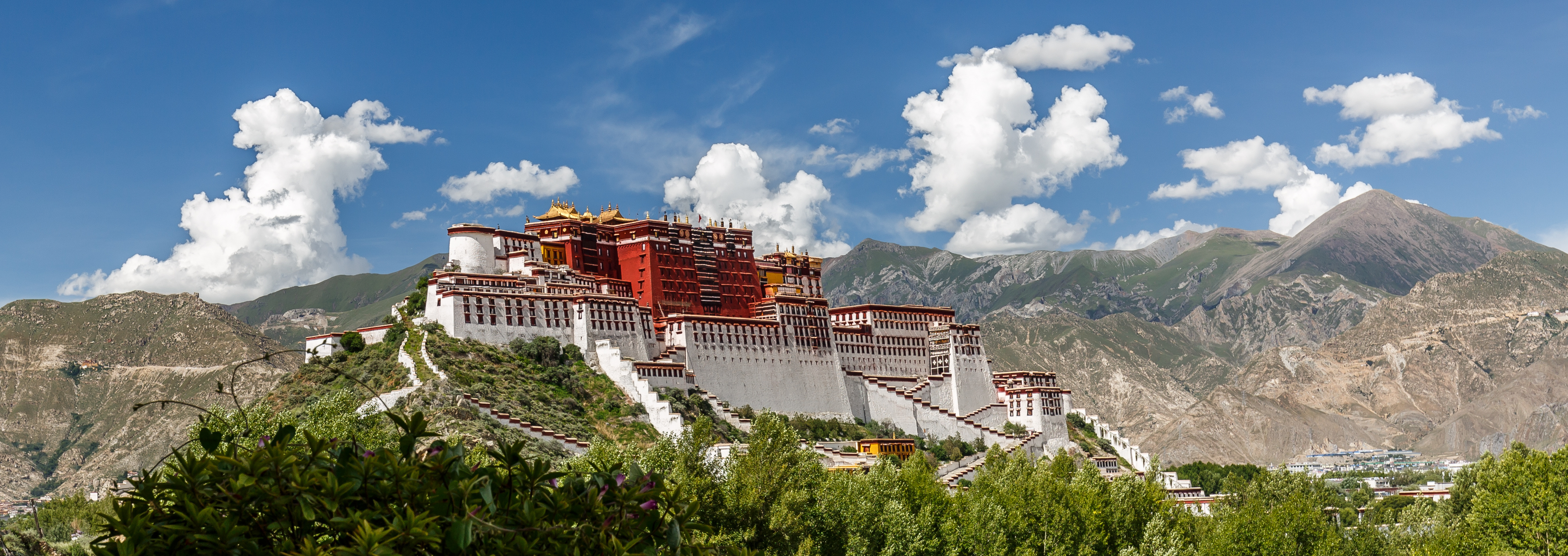 Potala Palace rises on its fortified hillside above Lhasa, red and white walls beneath a blue sky with mountains behind