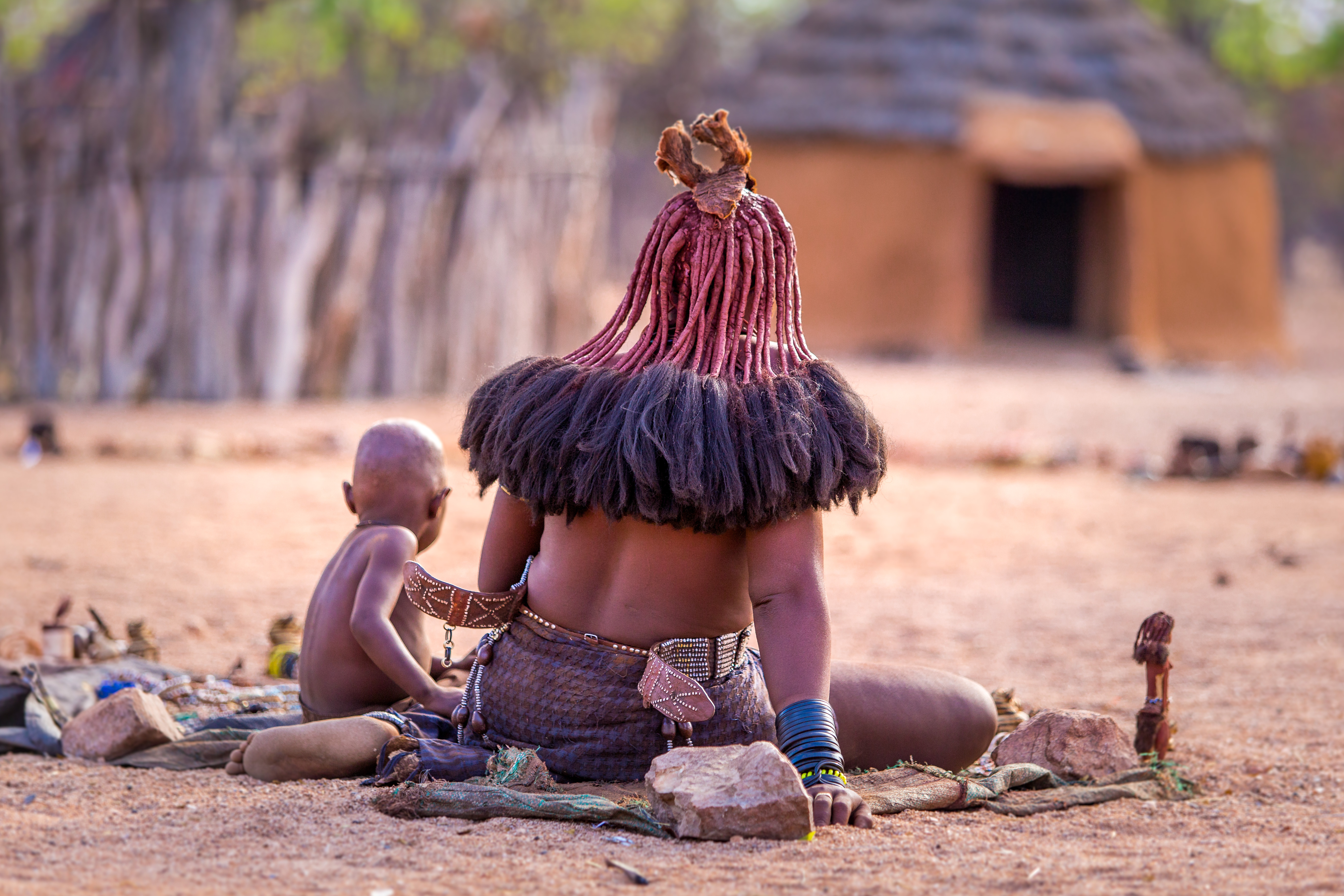 Himba woman with an ochre-dyed hairstyle and child sit on mats near a thatched-roof homestead in Namibia, both seen from behind