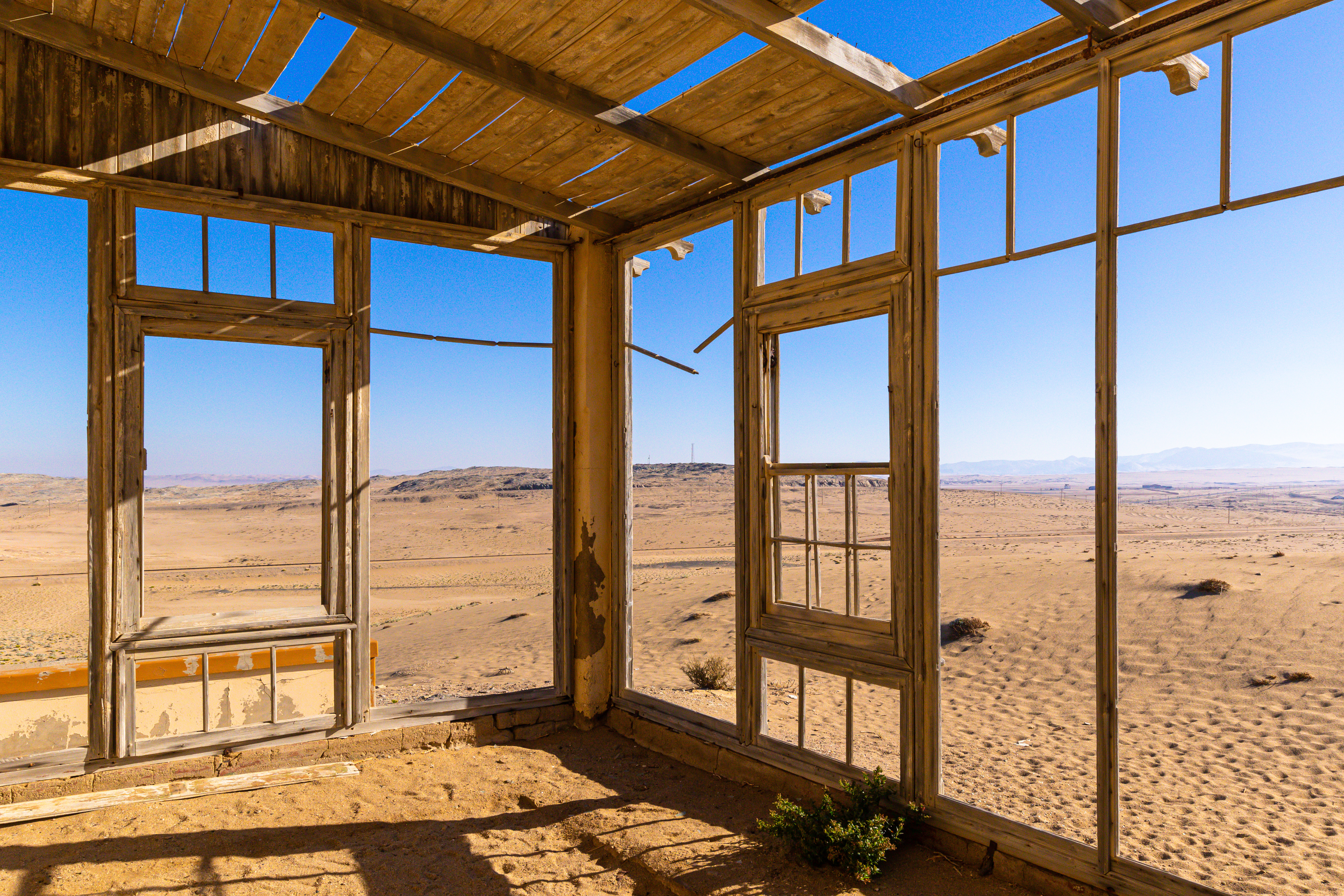 Sand drifts across the floor of a roofless, weathered building in Kolmanskop, with window frames opening onto desert dunes and blue sky